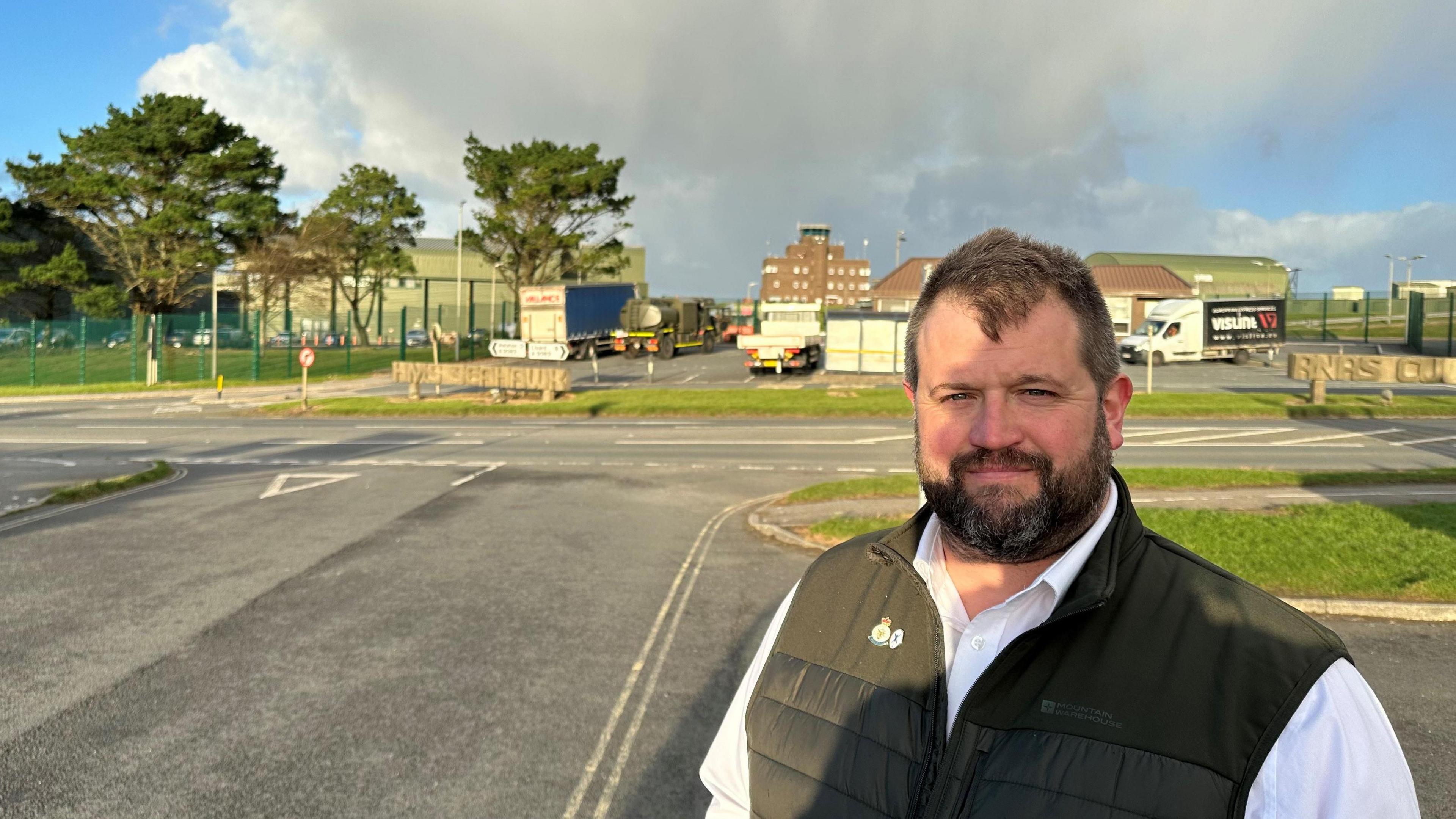 Daniel Thirkill stands outside the entrance to the air base. He has dark brown hair and a beard.