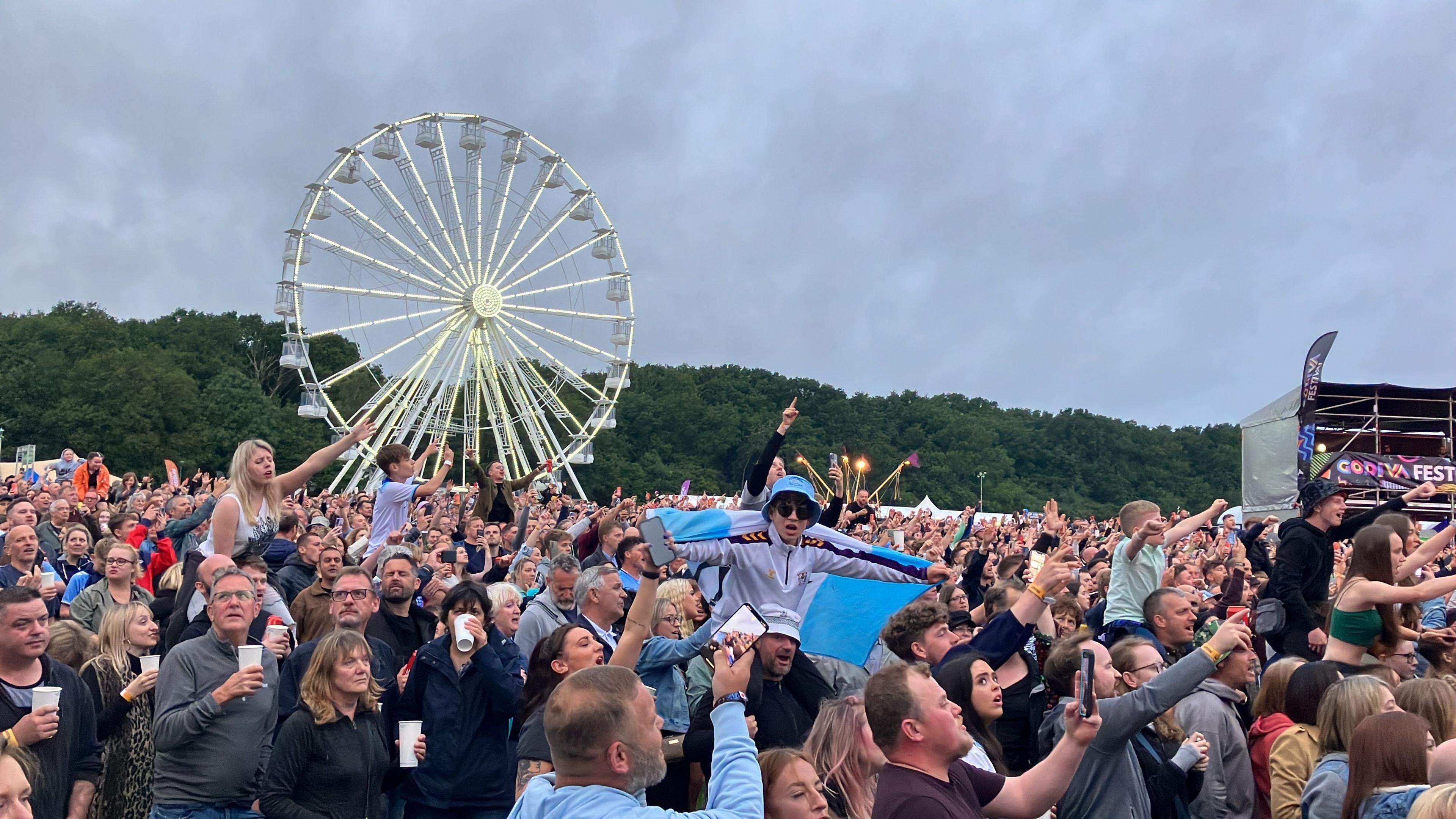 Hundreds of people in the Godiva Festival Crowd, with drinks, phones or hands in the air. A man is on another man's shoulders and is wearing a blue bucket hat and holding a blue flag. In the background is treeline and a Ferris wheel