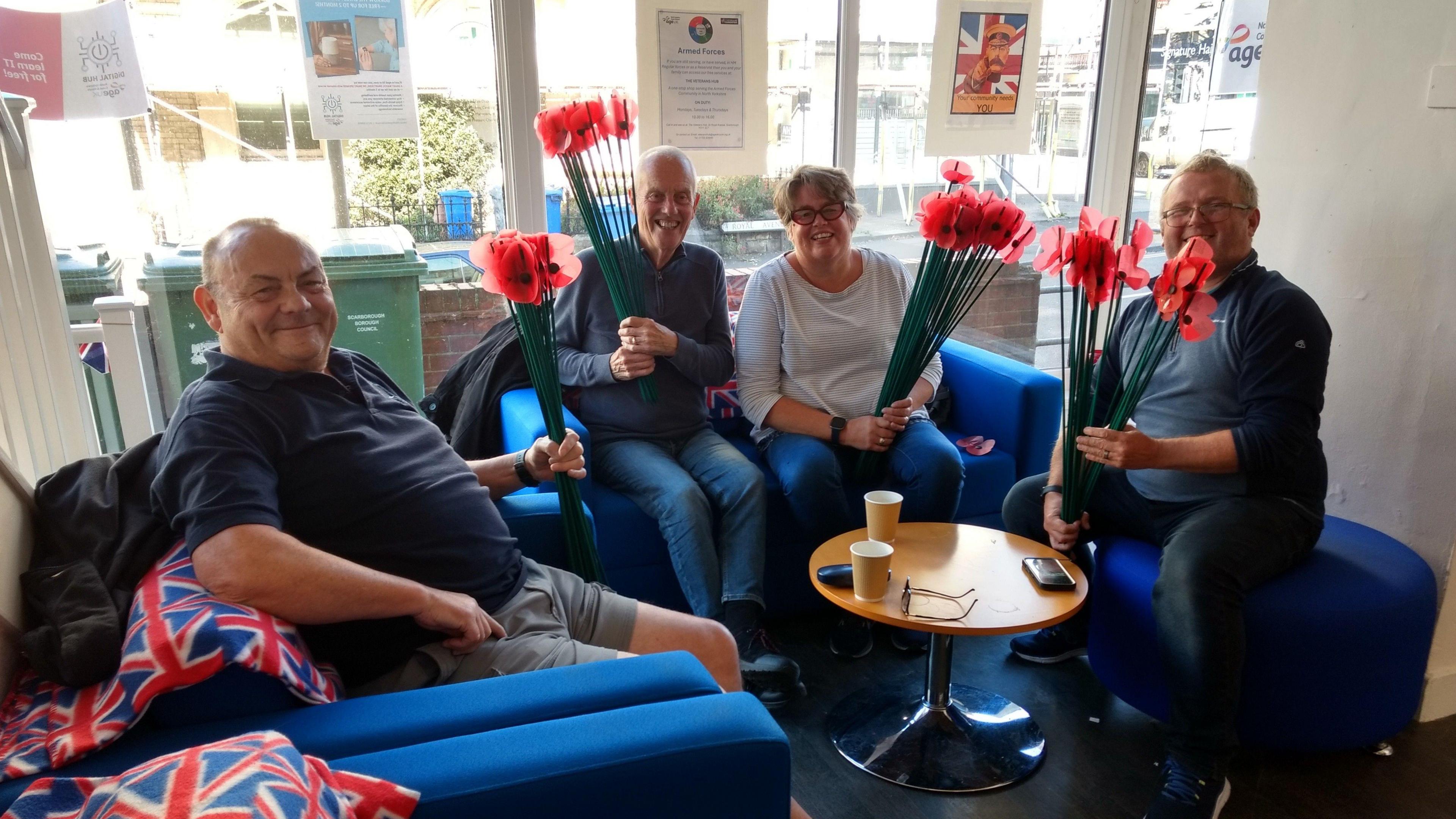 Four adults are sitting together in a bright, welcoming room. They’re gathered around a small, round wooden table, each seated on a blue chair. Each person is holding a bunch of red artificial poppies with green stems. Behind them, posters are visible on the windows.