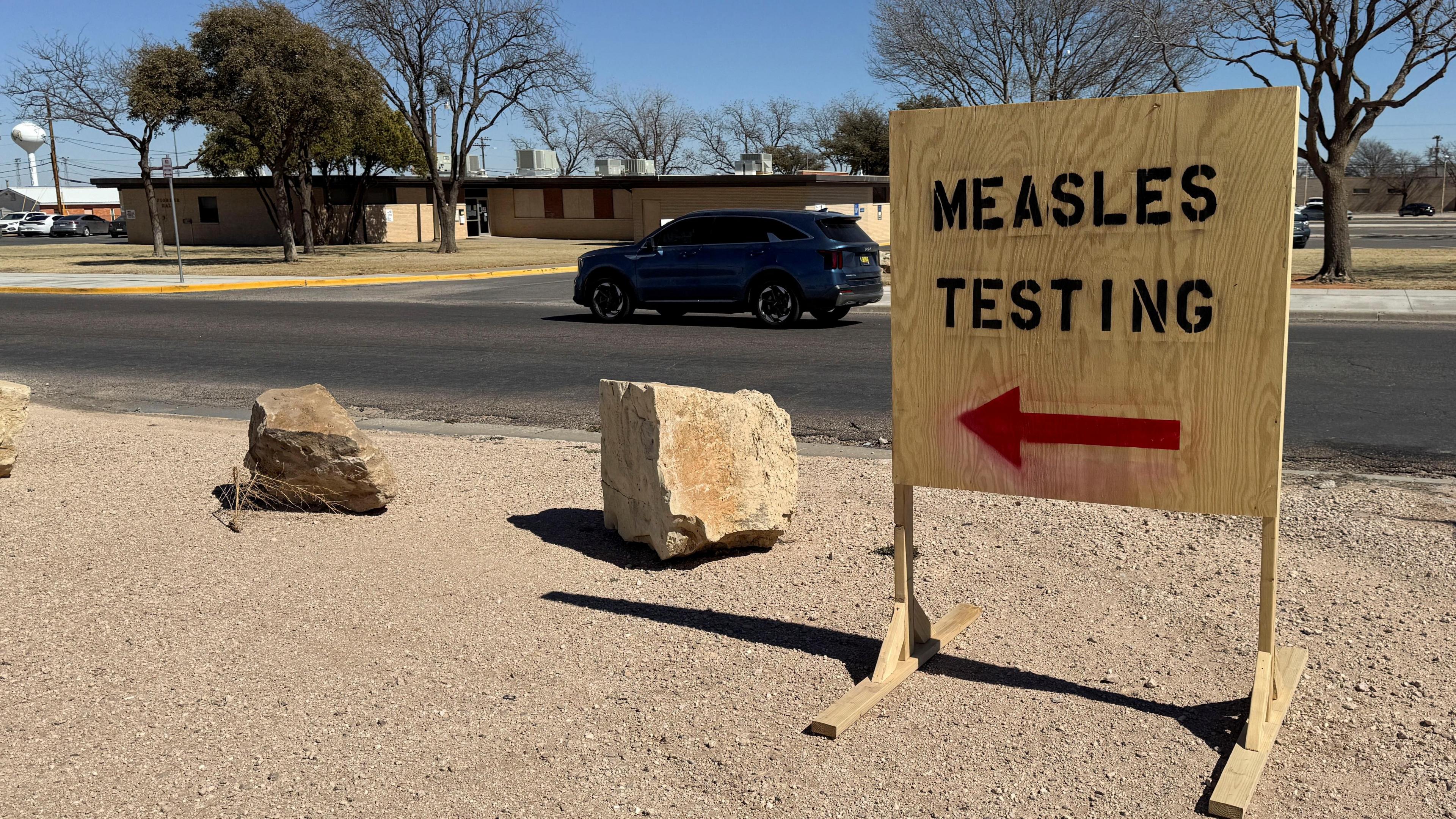 Plywood sign with black all-capital letters reading "Measles Testing" and a red arrow on sandy land in front of a street and squat building