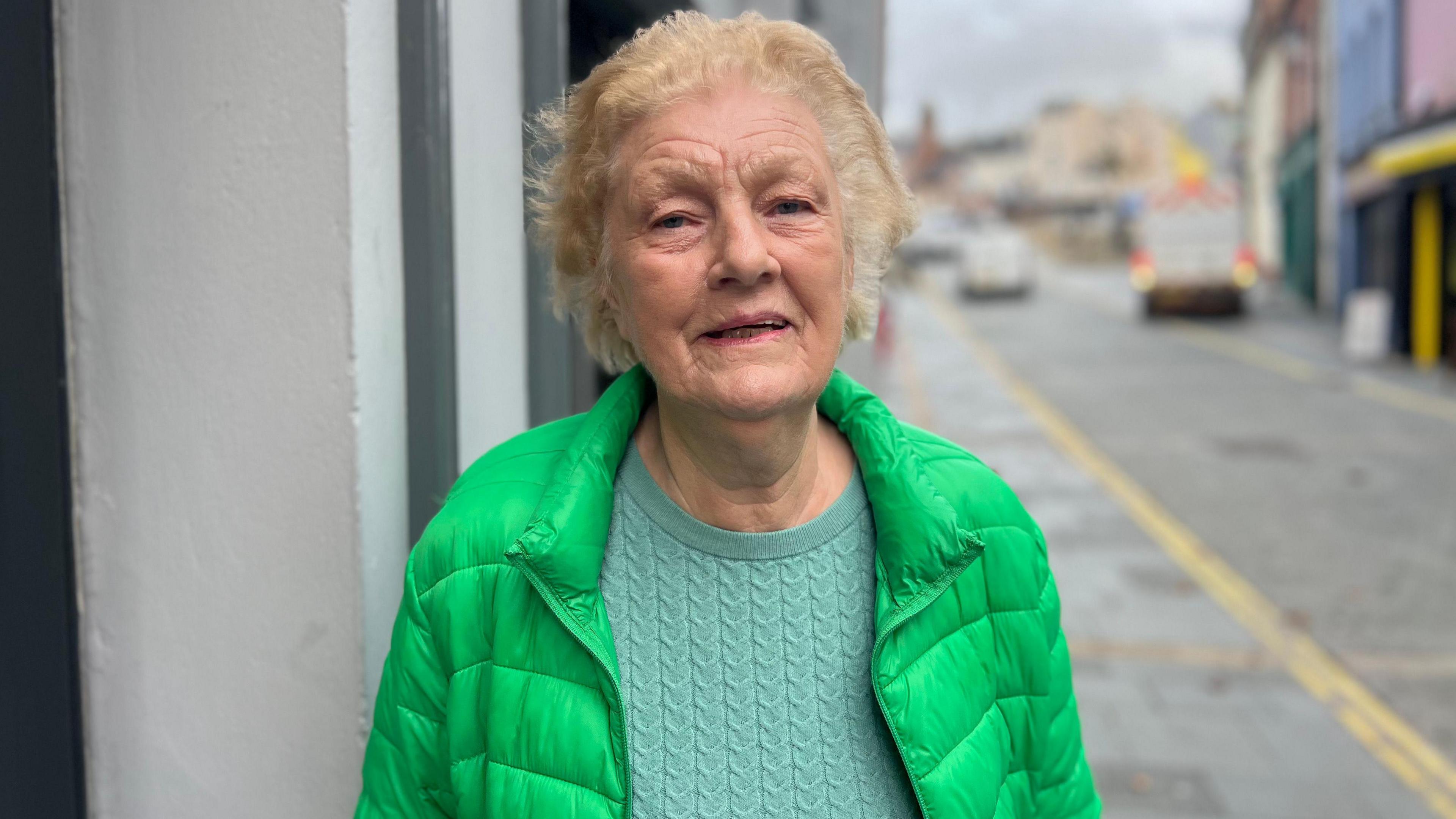 Veronica Doherty is standing in a street. She's wearing a green coat and a light green jumper. She has short hair.