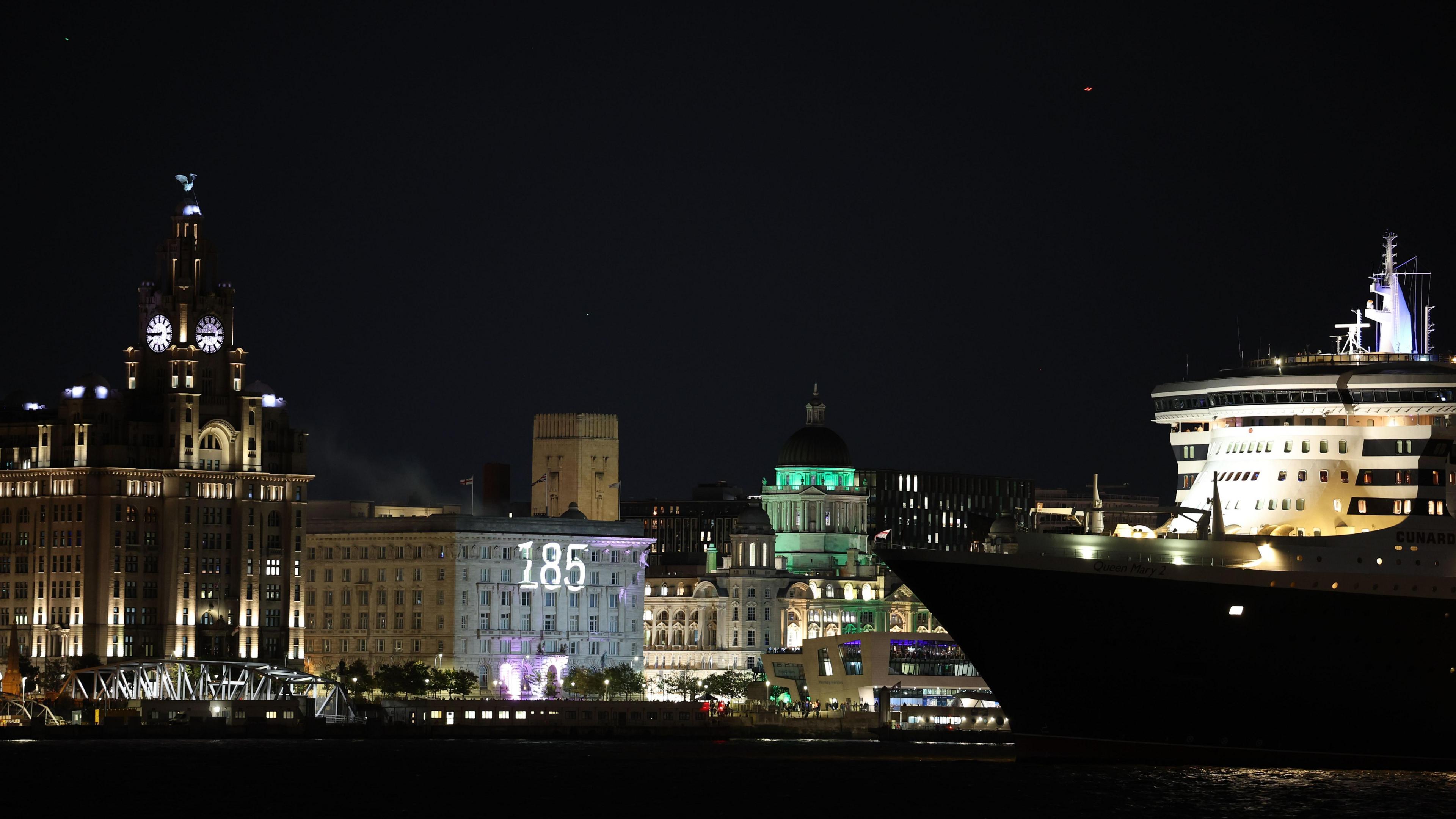The number 185 is projected on to the white Cunard building next to the Liver building on the Pier Head. The liner sails in front with its front white deck illuminated by light.