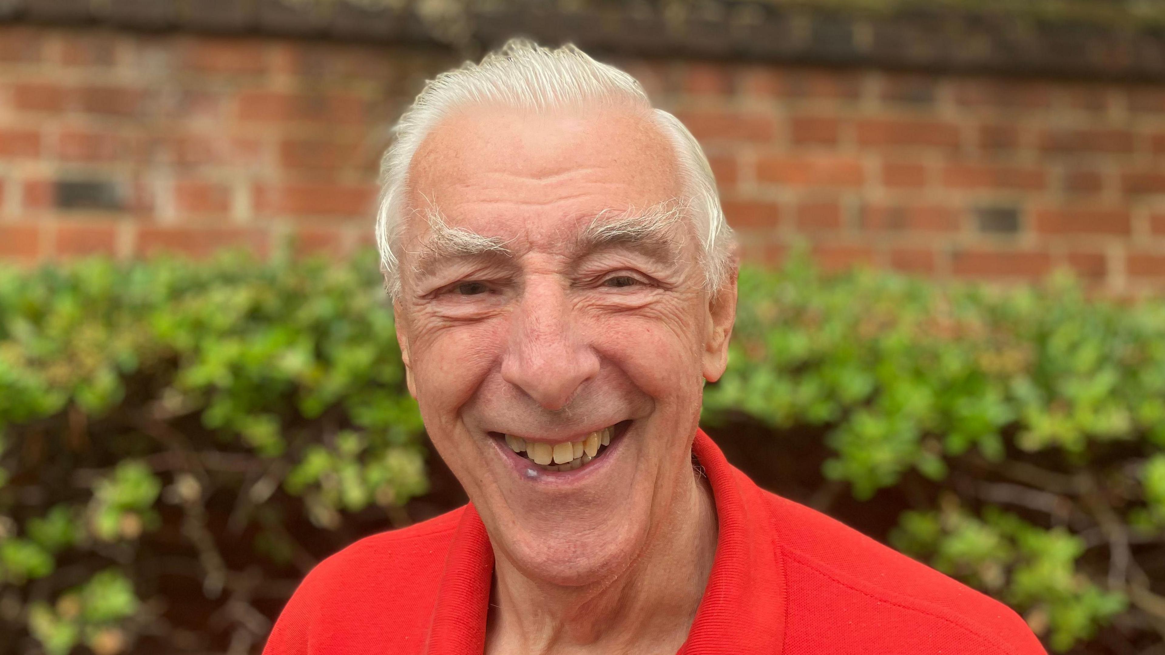 A close up view of Mr Read. He is smiling as he stands in front of a green bush and red brick wall. He has short grey hair and wears a red polo T-shirt.