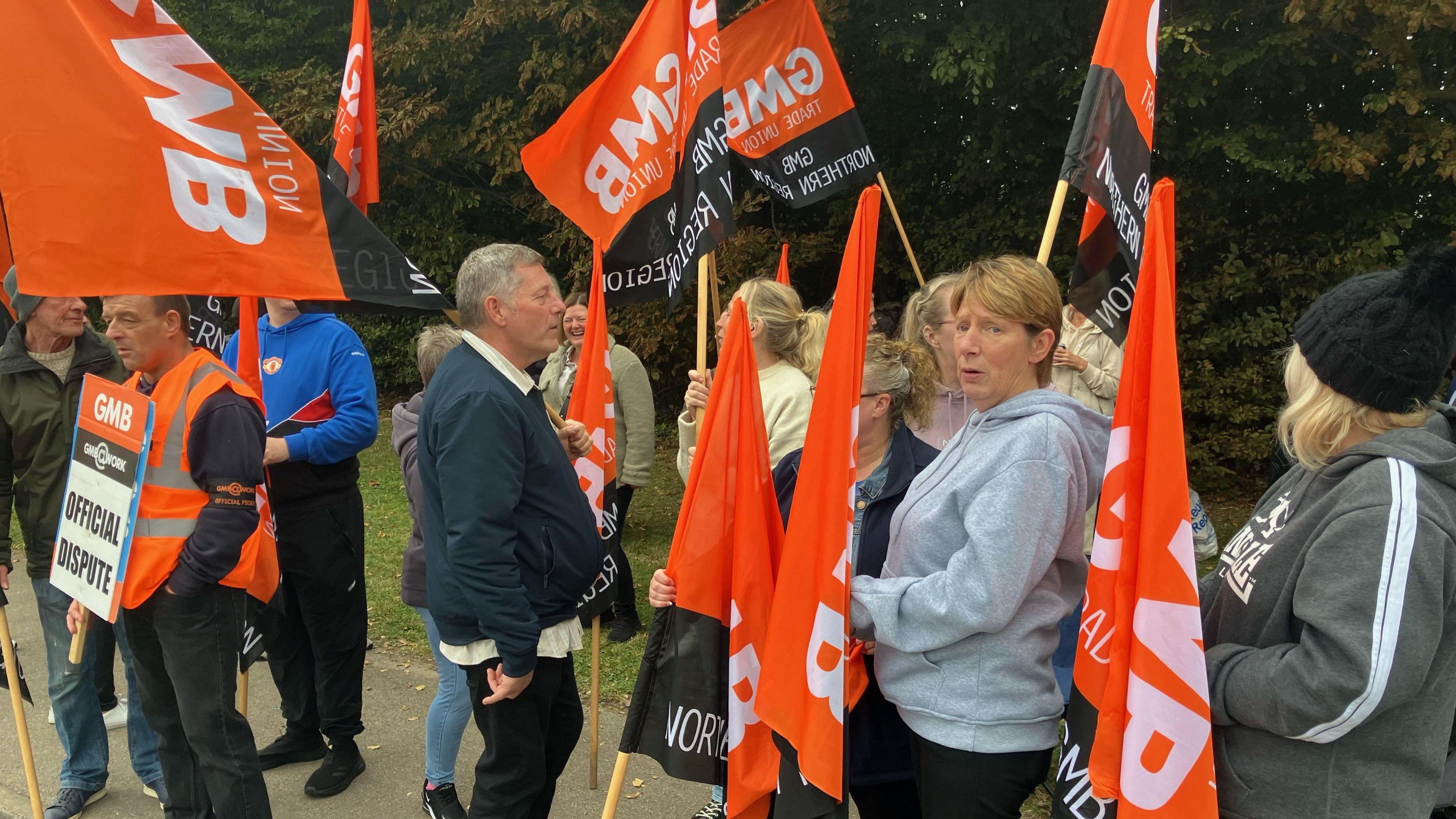 A dozen workers stand on a street holding orange and black "GMB" branded flags and signs. One of the signs reads: "OFFICIAL DISPUTE".