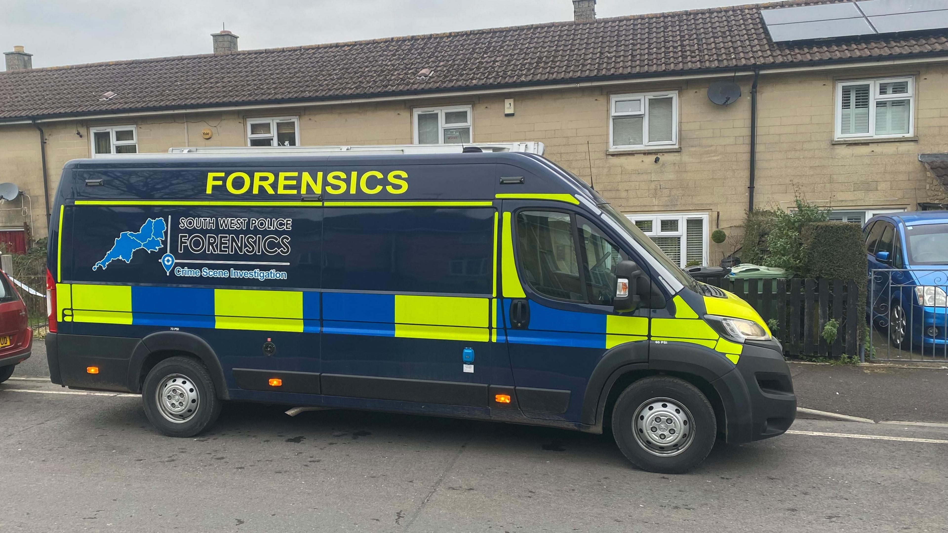 A big, navy police van saying 'South West Police Forensics' on it, parked outside a property.