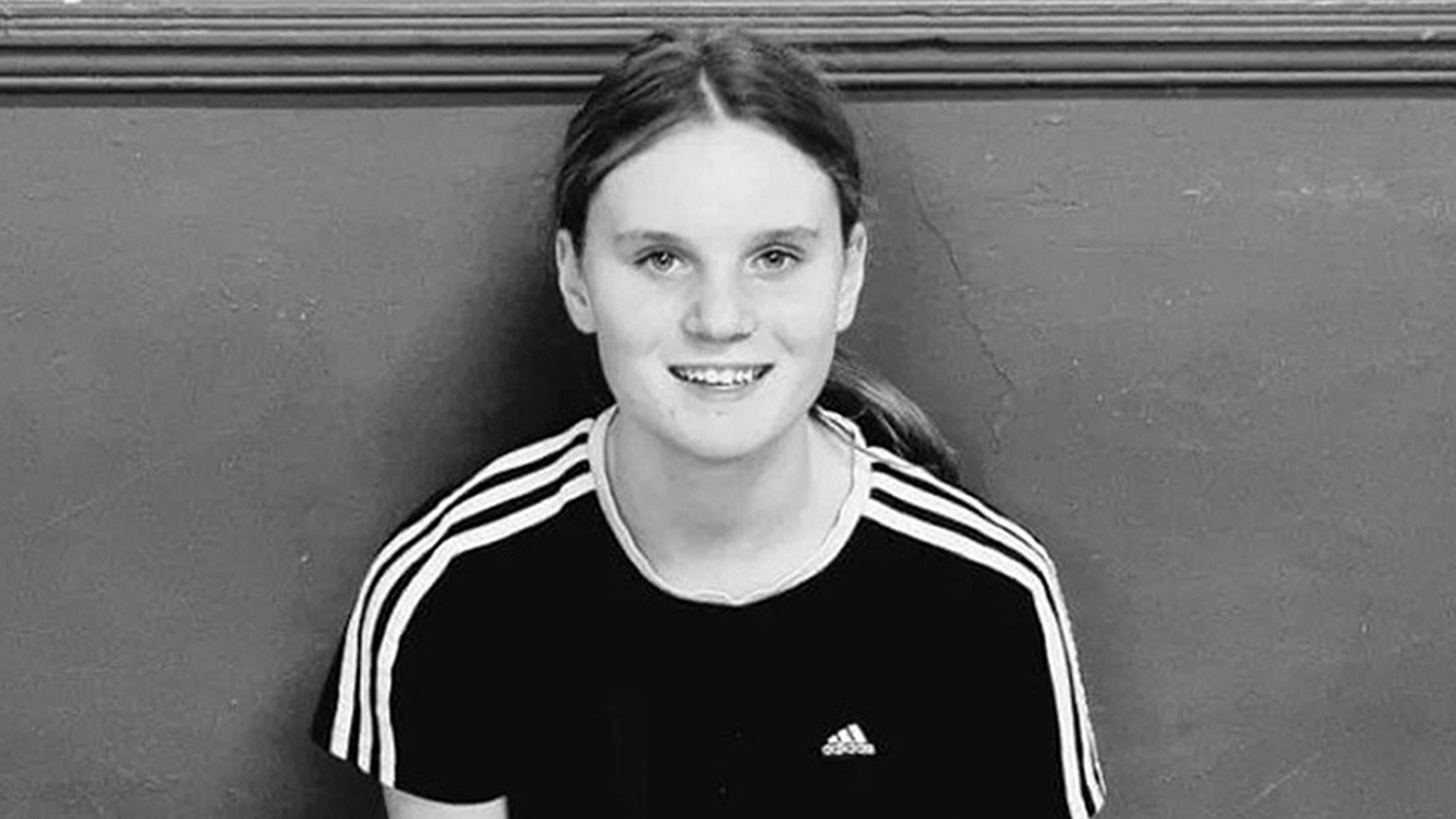 Black and white picture of Holly. She has long brown hair pulled back into a pony tail and is wearing a black Adidas t-shirt with three white stripes down each arm. She is sitting with her back against a wall and is smiling at the camera