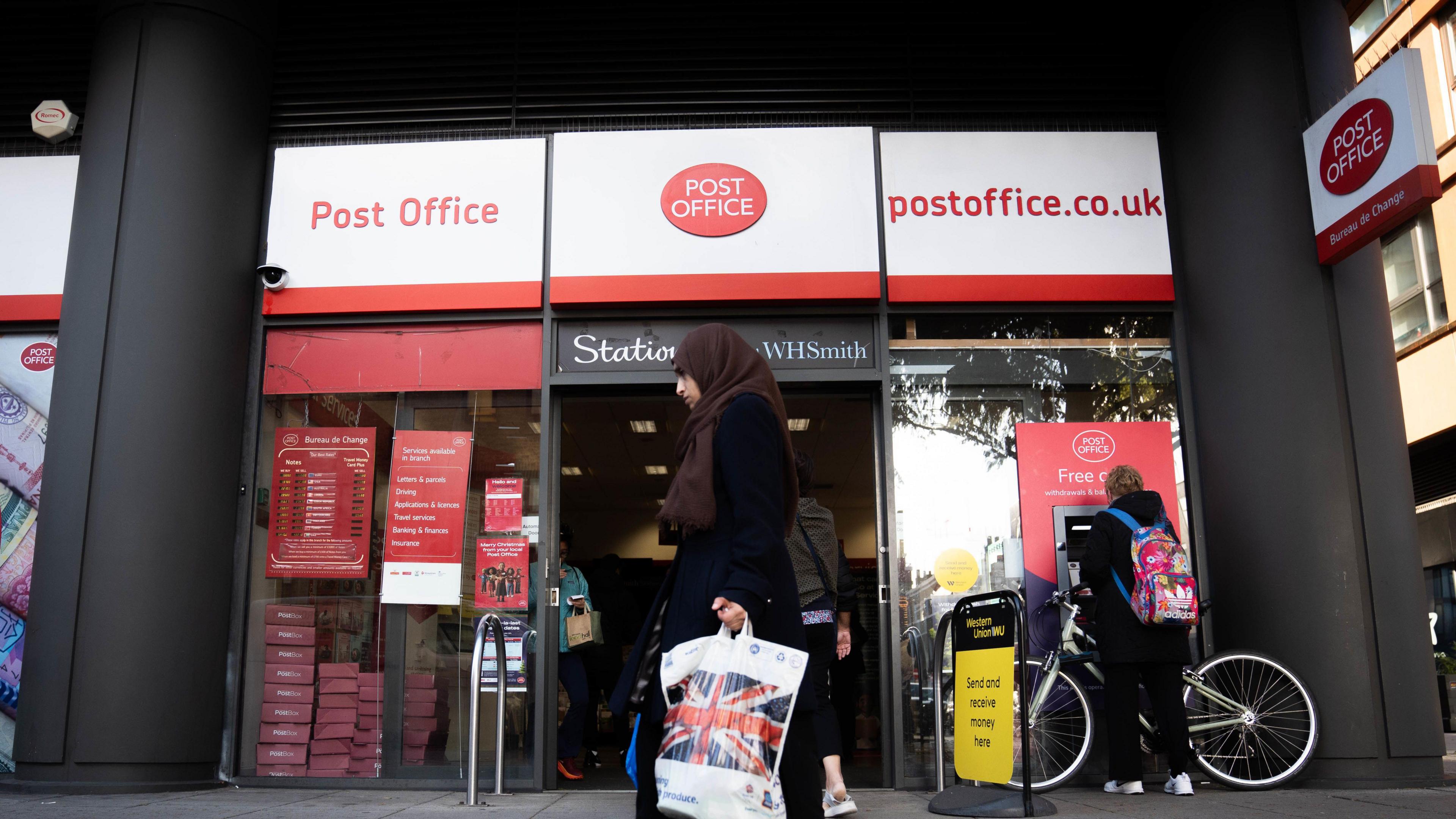 A woman walks with shopping bag past a Post Office branch on a high street