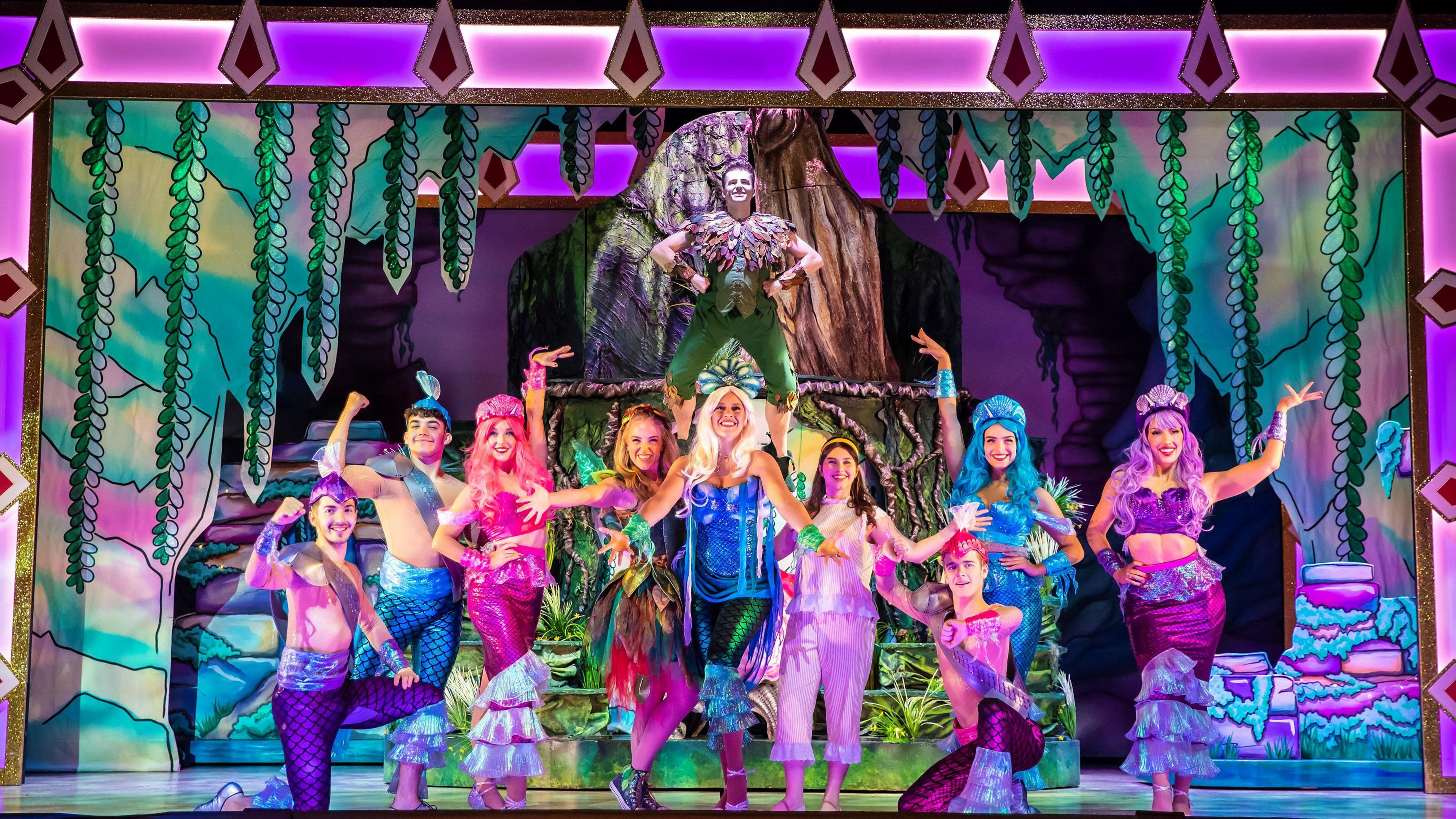 The cast of Peter Pan on stage, showing a colourful scene of dancers and performers, dressed like mermaids and mermen, in deep purple colours. Peter Pan is behind them flying with his hands on his hips. 