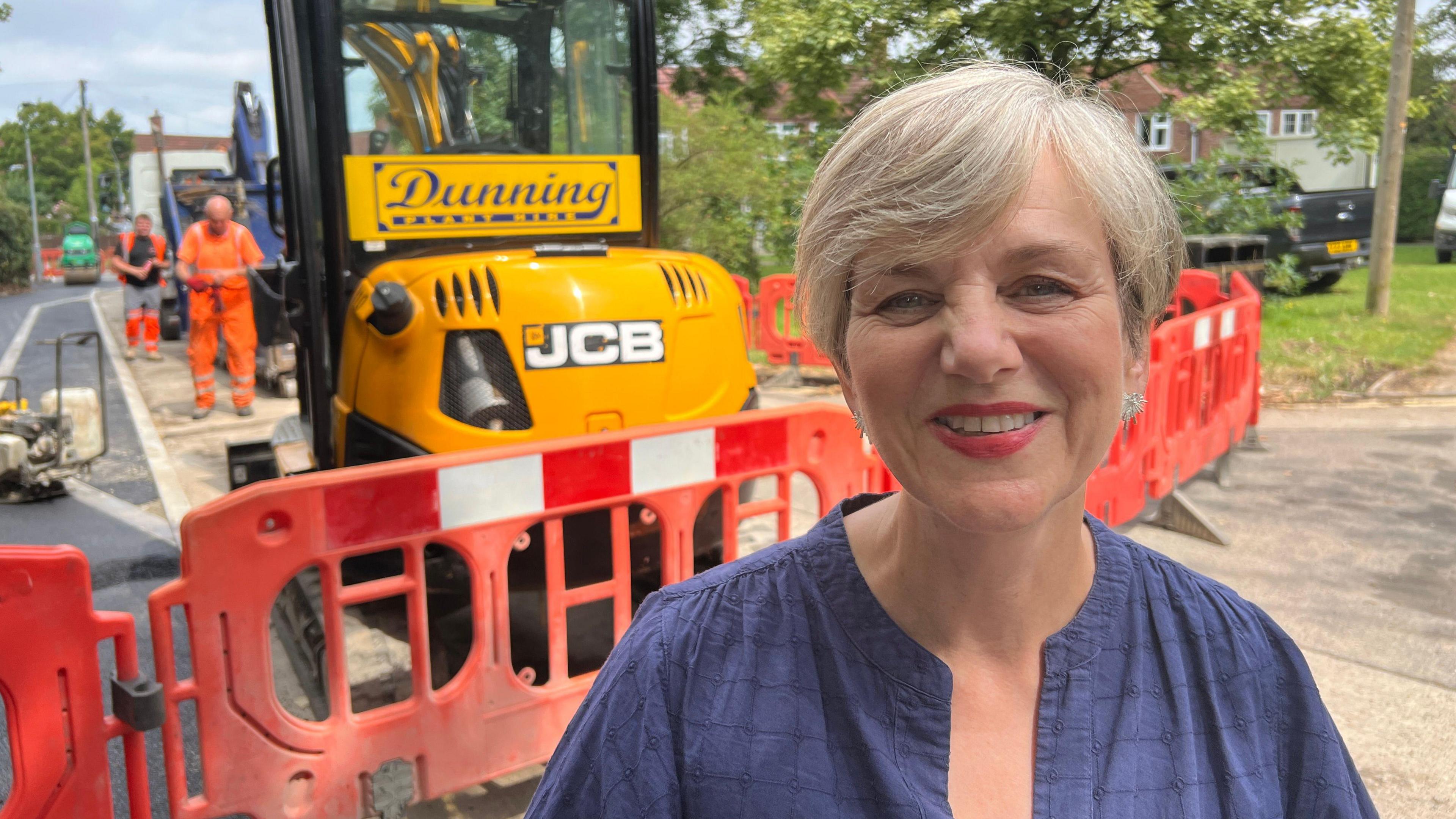 Roads minister Lilian Greenwood is pictured smiling in a blue top stood in front of a JCB and road resurfacing works