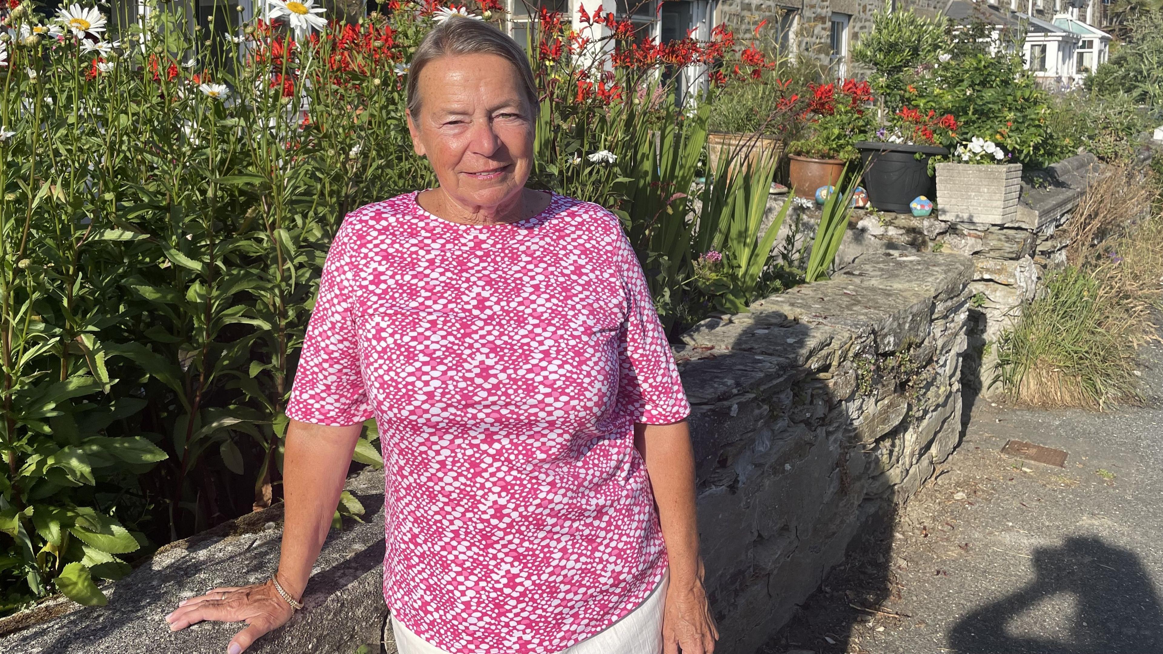 A woman in a red and white top leans on a wall in front of a flower bed