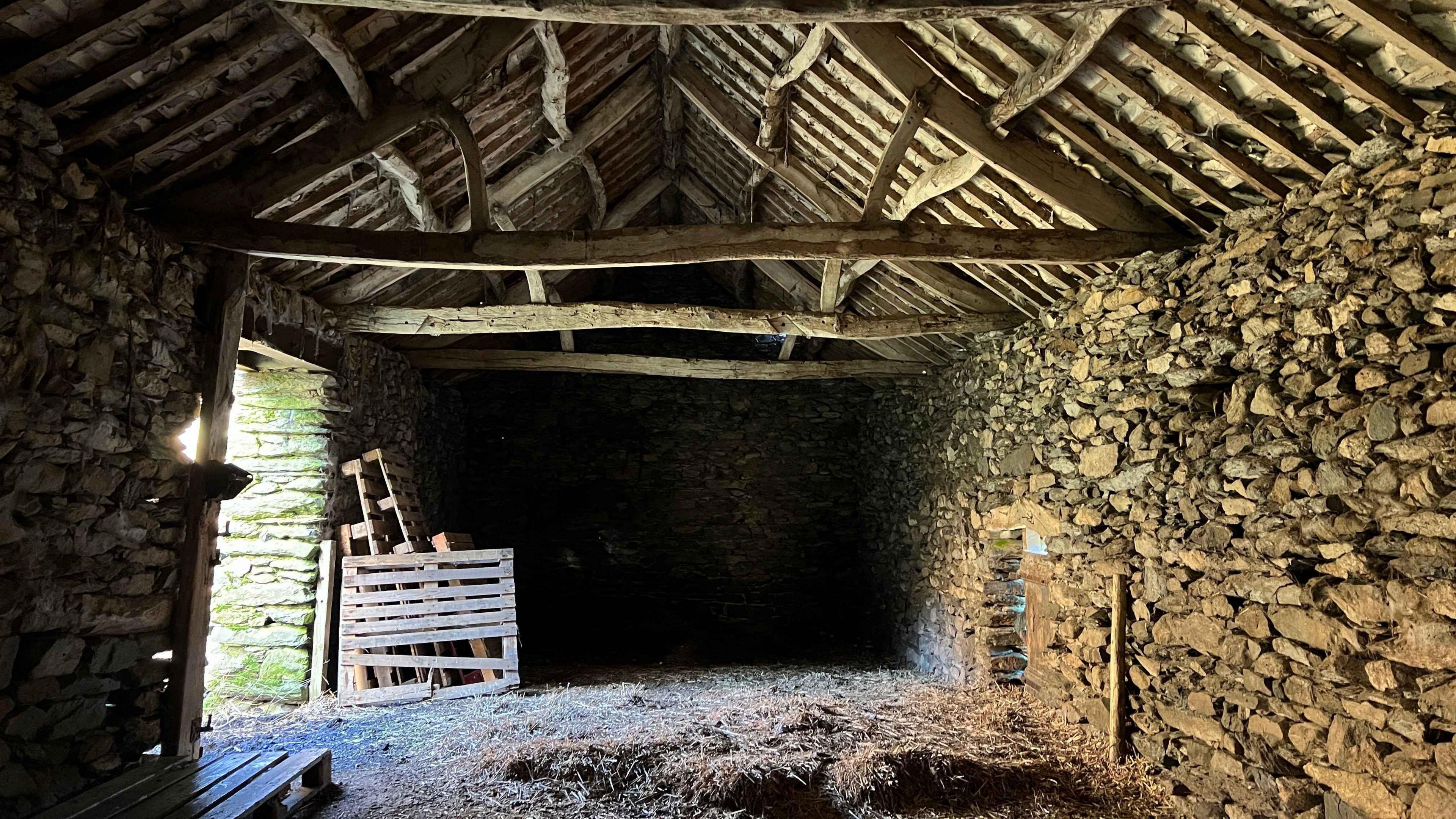 Inside of old rural barn with wooden roof with beams. The walls are made of irregularly shaped stones. There is a wooden pallet by the entrance which is letting in sunshine into the barn. The floor is covered in hay. 