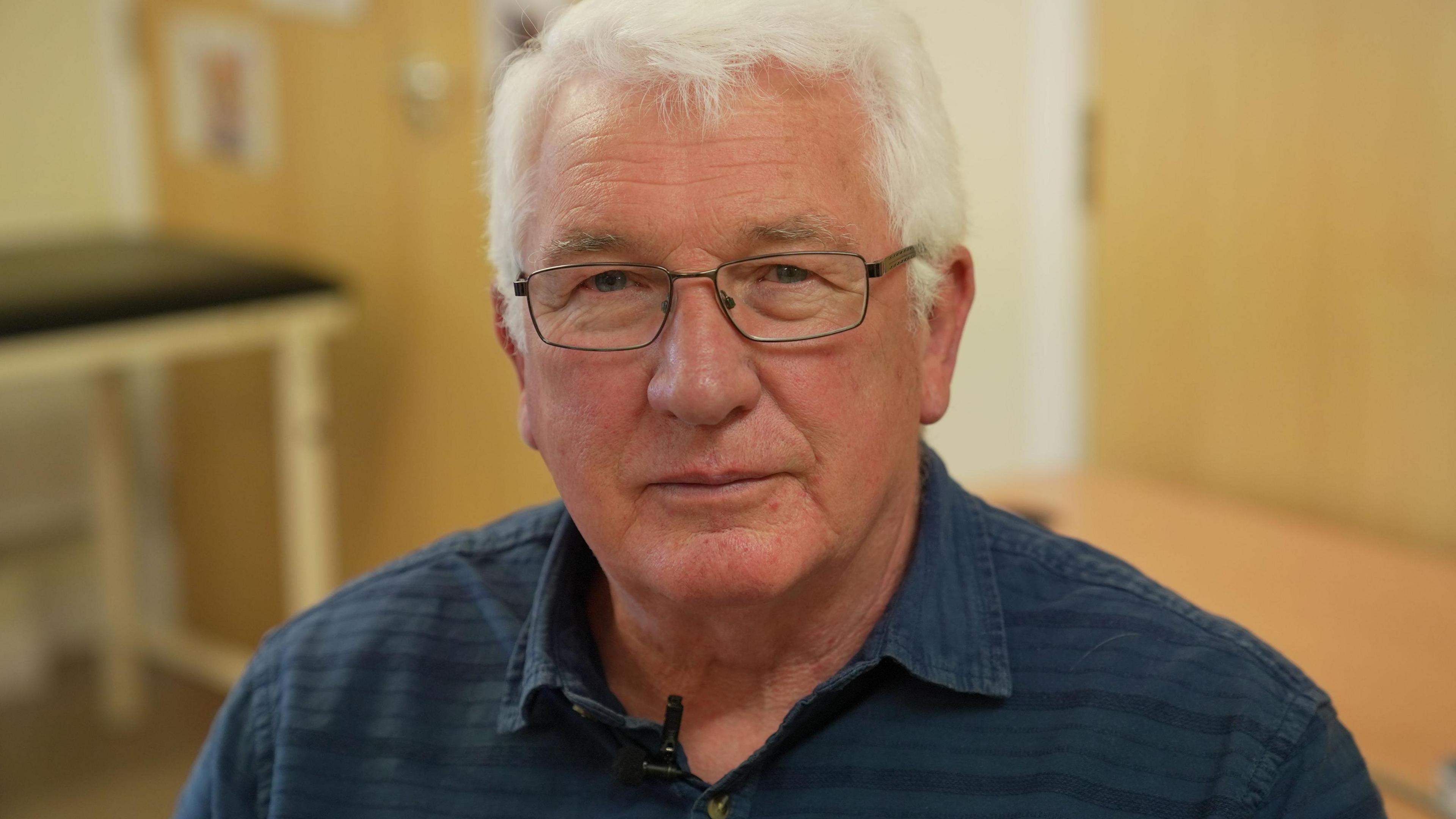 Ged Cashley is wearing a blue shirt and glasses.  The 72 year old suffered a cardiac arrest in 2018 and now has annual health checks.