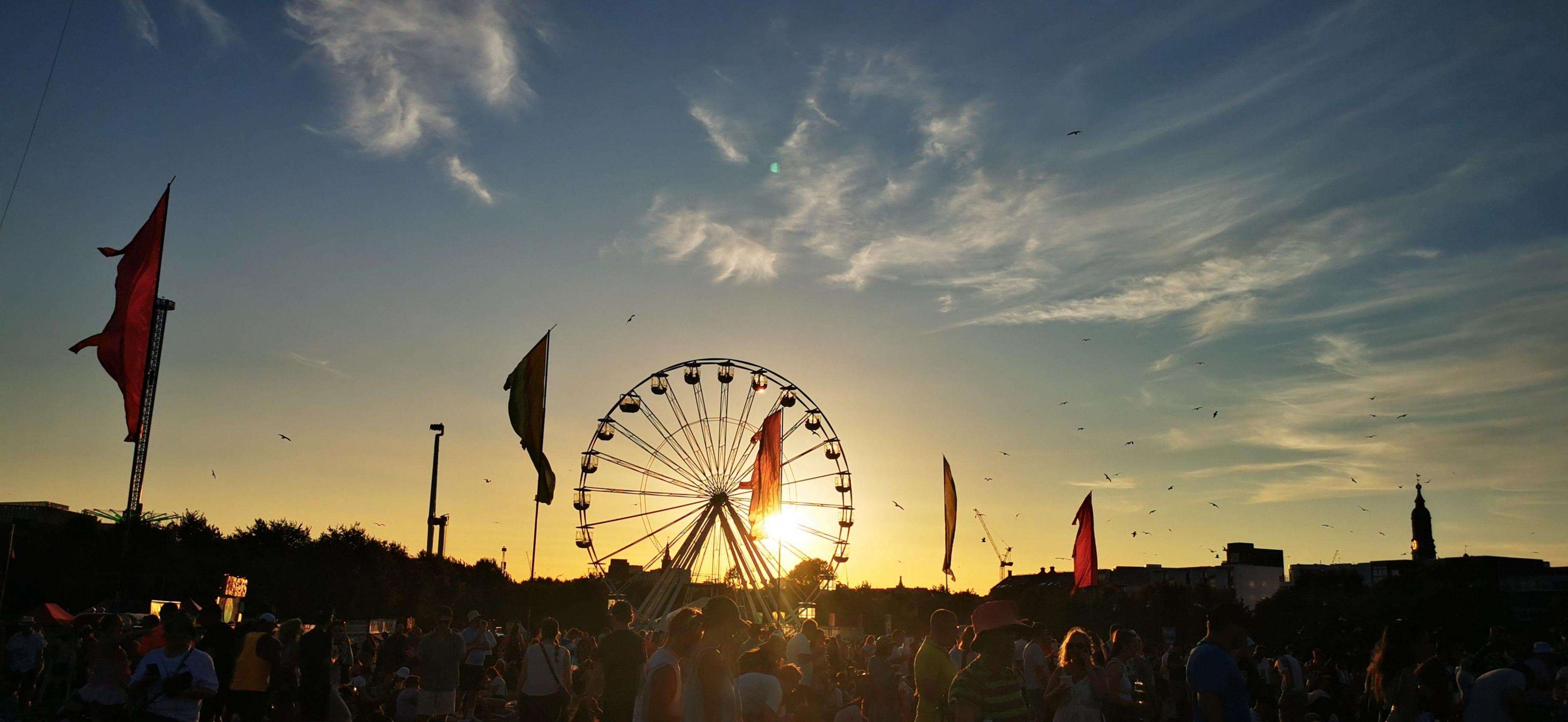 The sun slowly setting over a music festival crowd. There is a large ferris wheel and flags