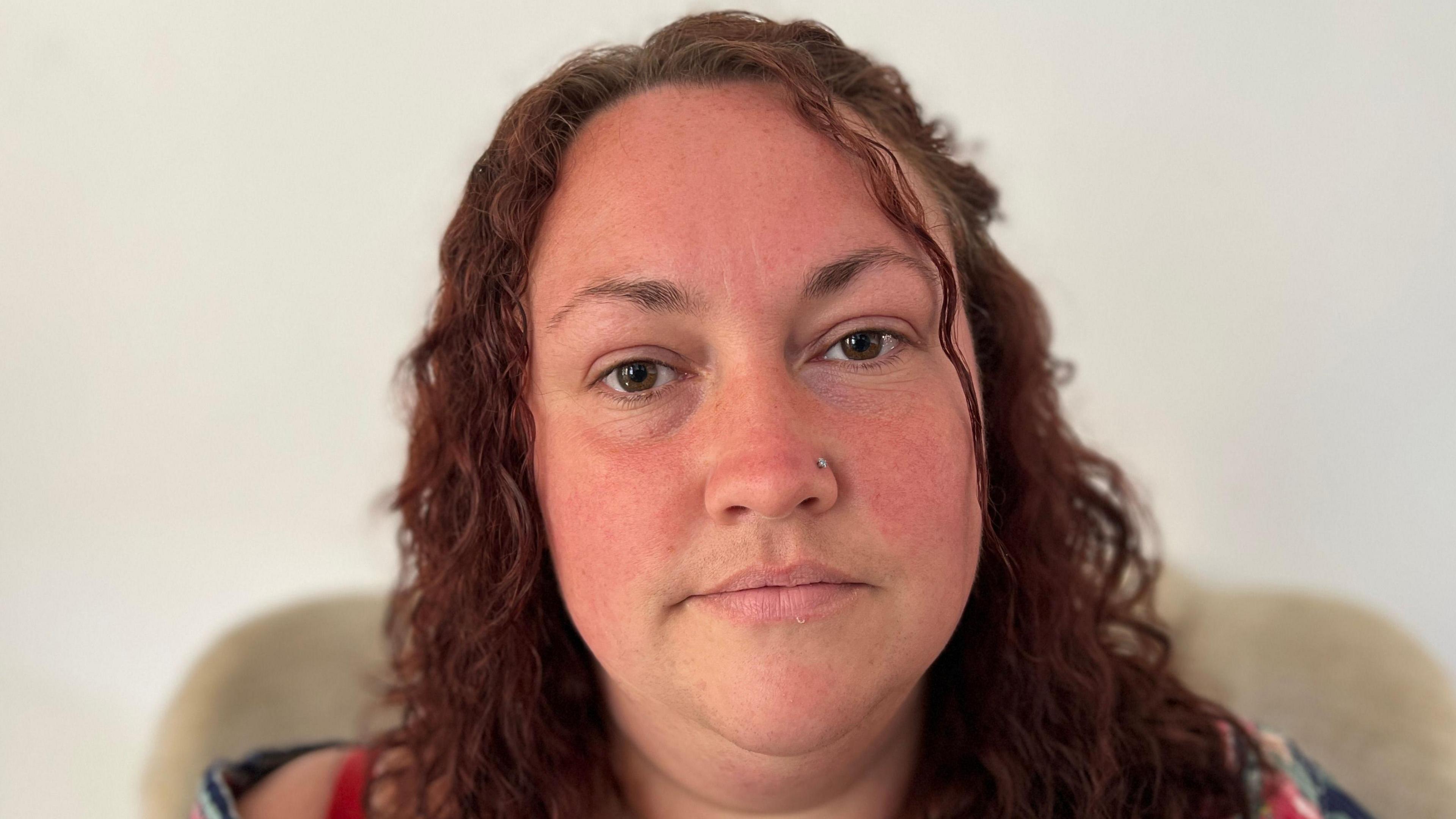 A woman with curly brown hair, brown eyes and a nose piercing. She is sitting on a chair against a white background.