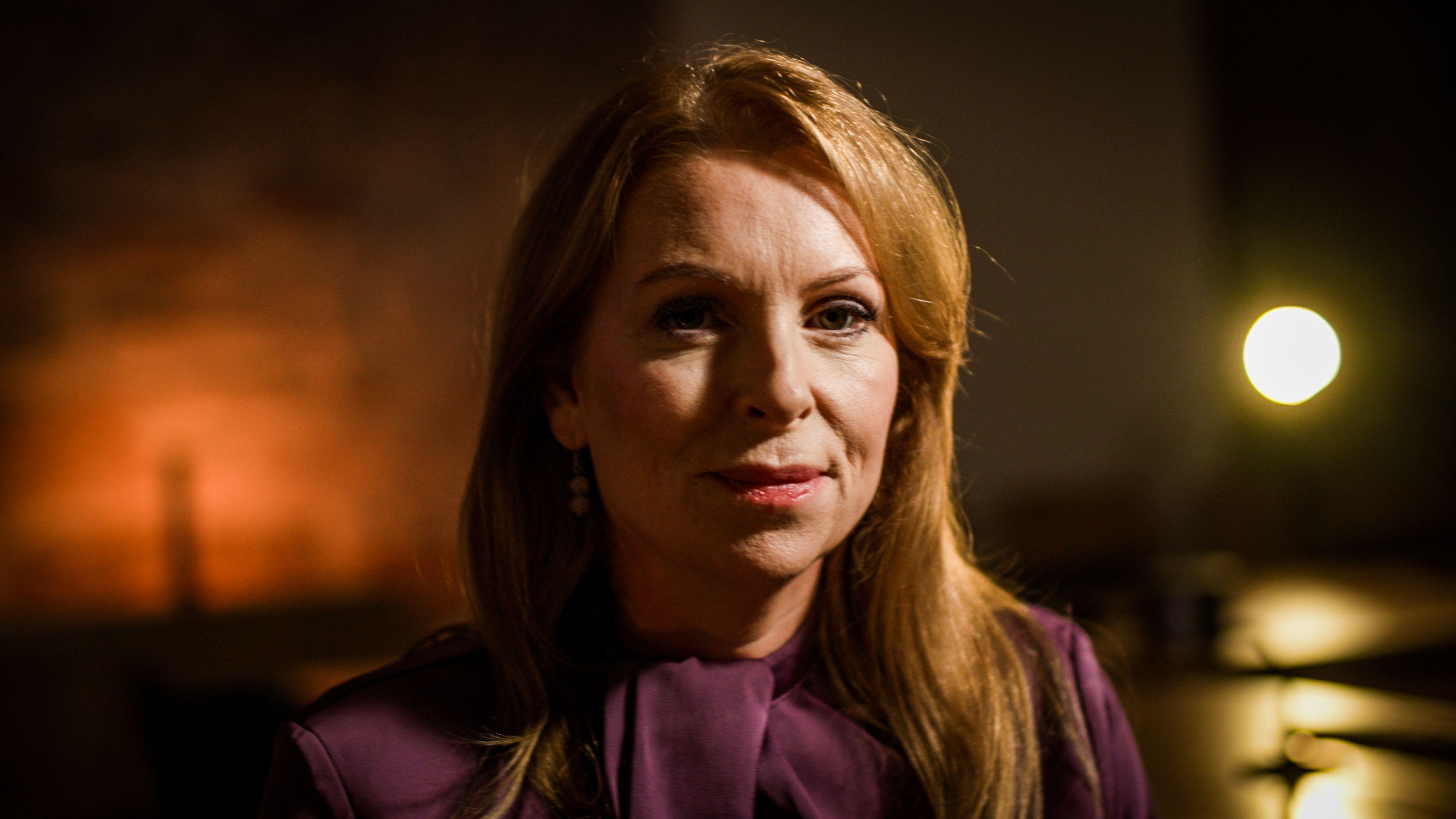 Ash Regan has long, red hair with a side parting. She is wearing a deep purple top featuring a high neckline with a bow at the front. The setting is dimly lit with warm tones. A light is shining behind her, casting a soft glow, while the rest of the background is blurred.