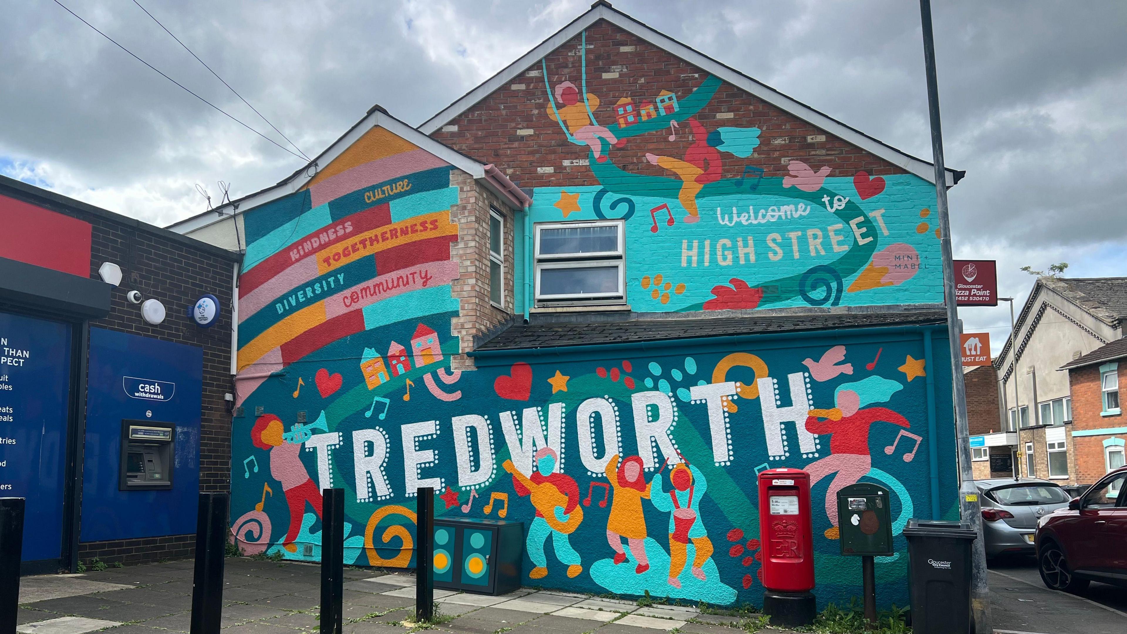 The side of a building which has been painted. Tredworth is painted along the bottom, with silhouettes of people dancing and playing musical instruments. The top of the building says 'welcome to High Street'.