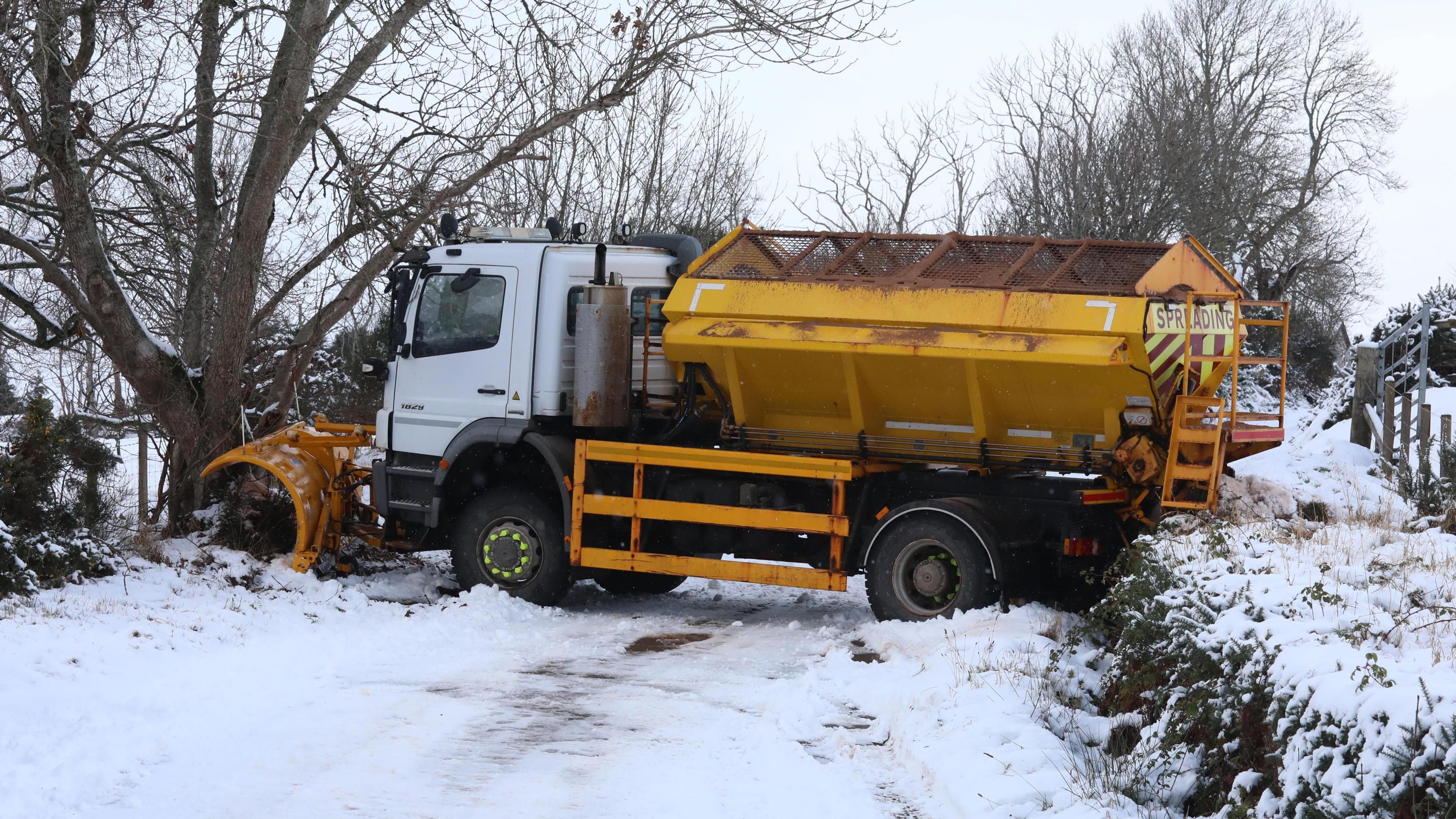 Snow plough stuck on snow-covered road. The vehicle is across the width of the road, wedged between a tree and a grass verge 