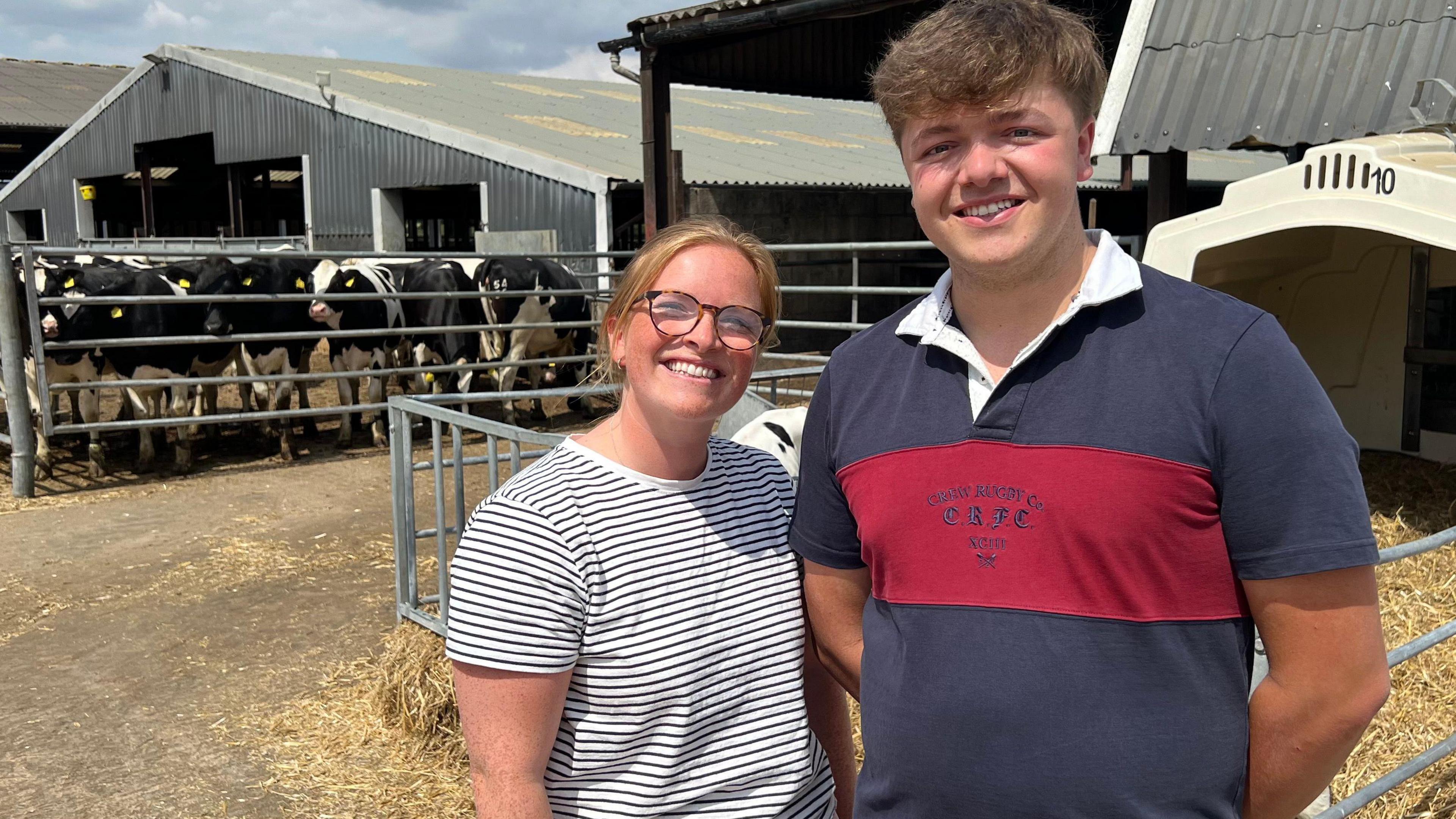 Lara (left) is wearing a blue and white striped tee shirt and has tied-back blonde hair and glasses. She is standing next to Sam and smiling at the camera. Next to her is Sam in a navy and red short sleeved rugby shirt, he has short brown hair. In the background is a farmyard and barn with cows standing by a gate.