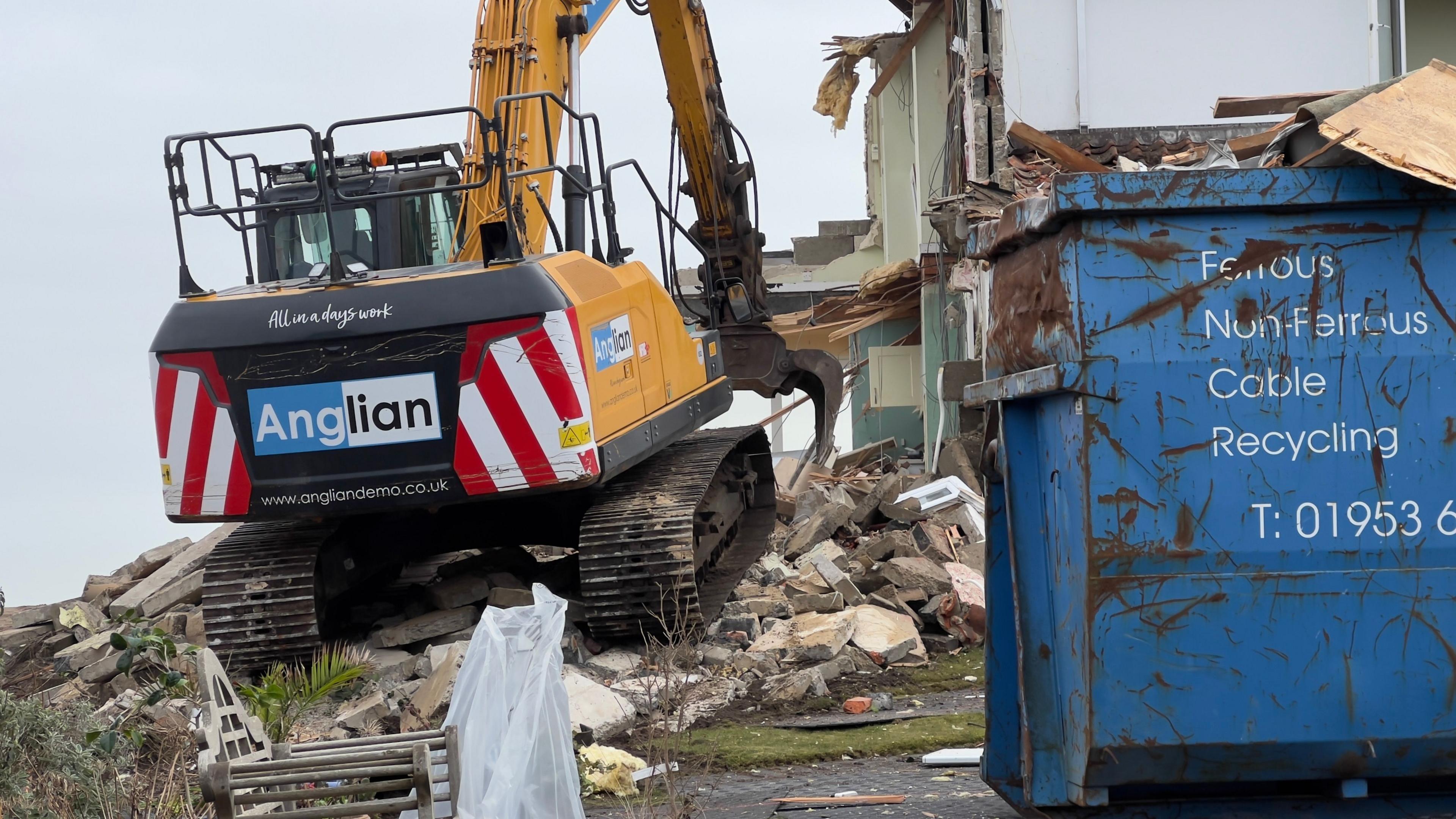 A close-up of a digger demolishing a home. A large industrial blue skip is on the right. Rubble and debris from the home is scattered along the ground and underneath the digger.