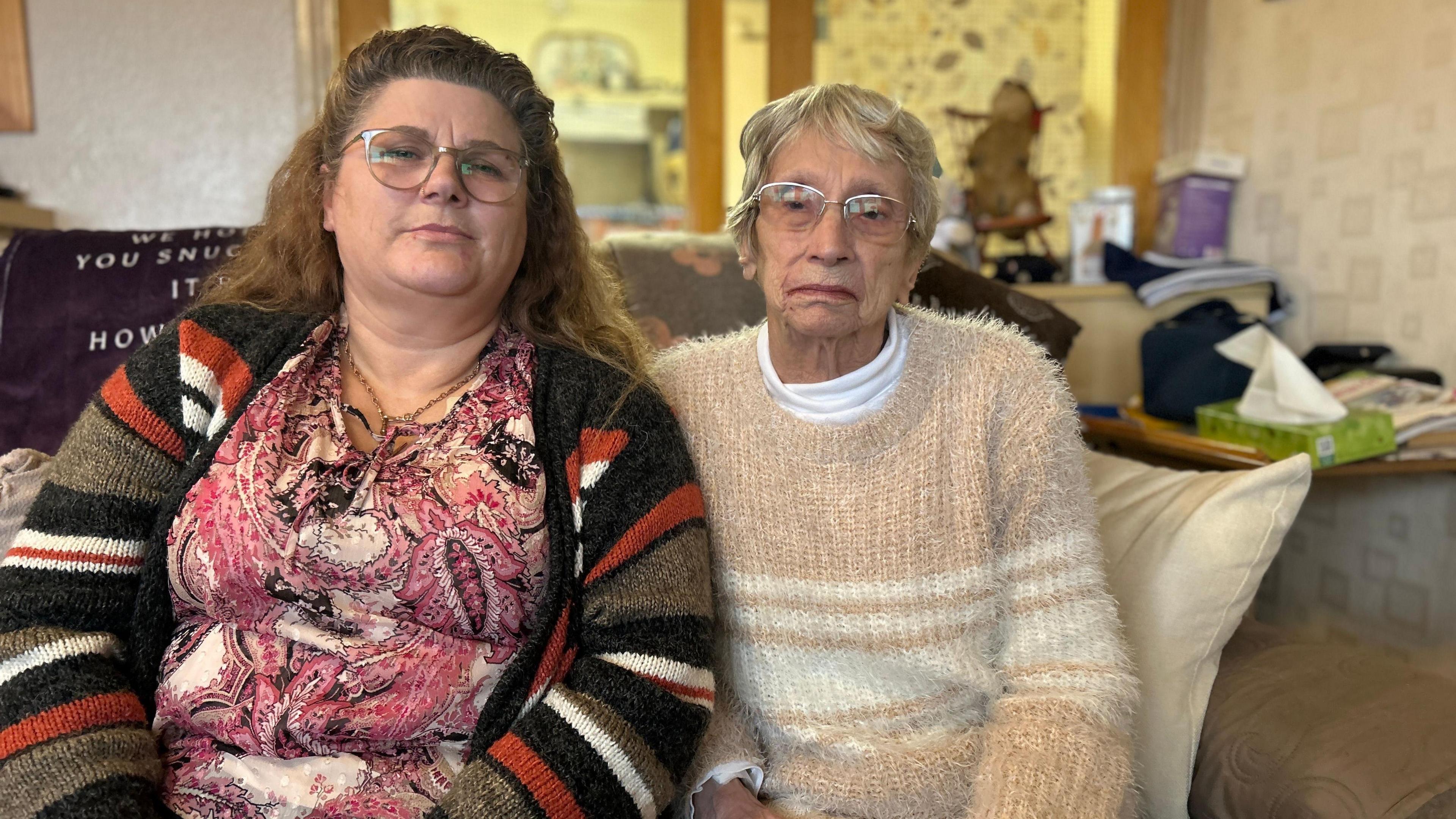 Lois Dehan with her mother Pamela, who is elderly. They are both wearing glasses and posing for a picture. They are wearing warm clothes and sitting on a sofa.