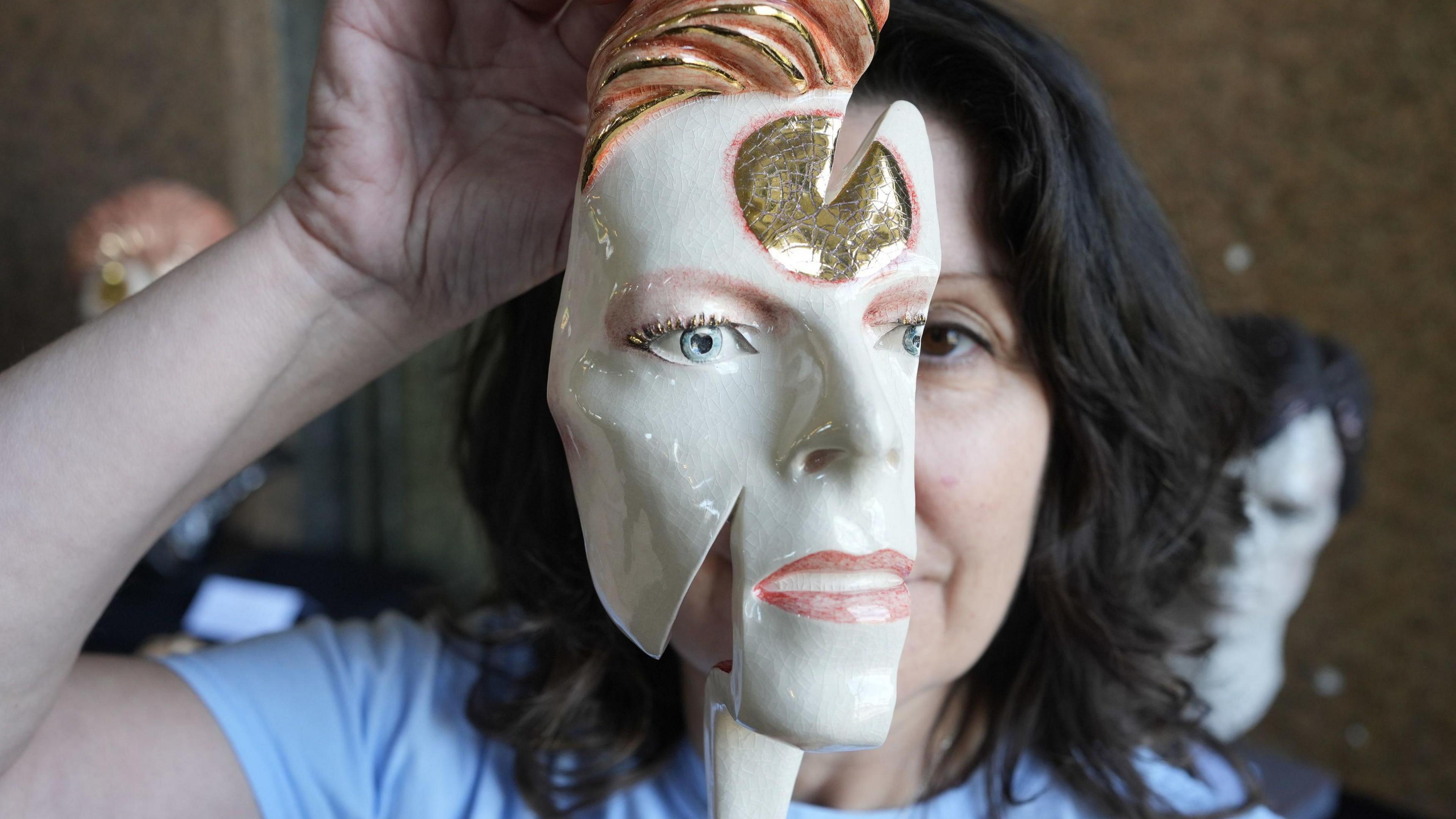 A woman with black hair and a white T-shirt holds a white, gold and peach-coloured mask of David Bowie's Aladdin Sane character in front of her face, covering half of it