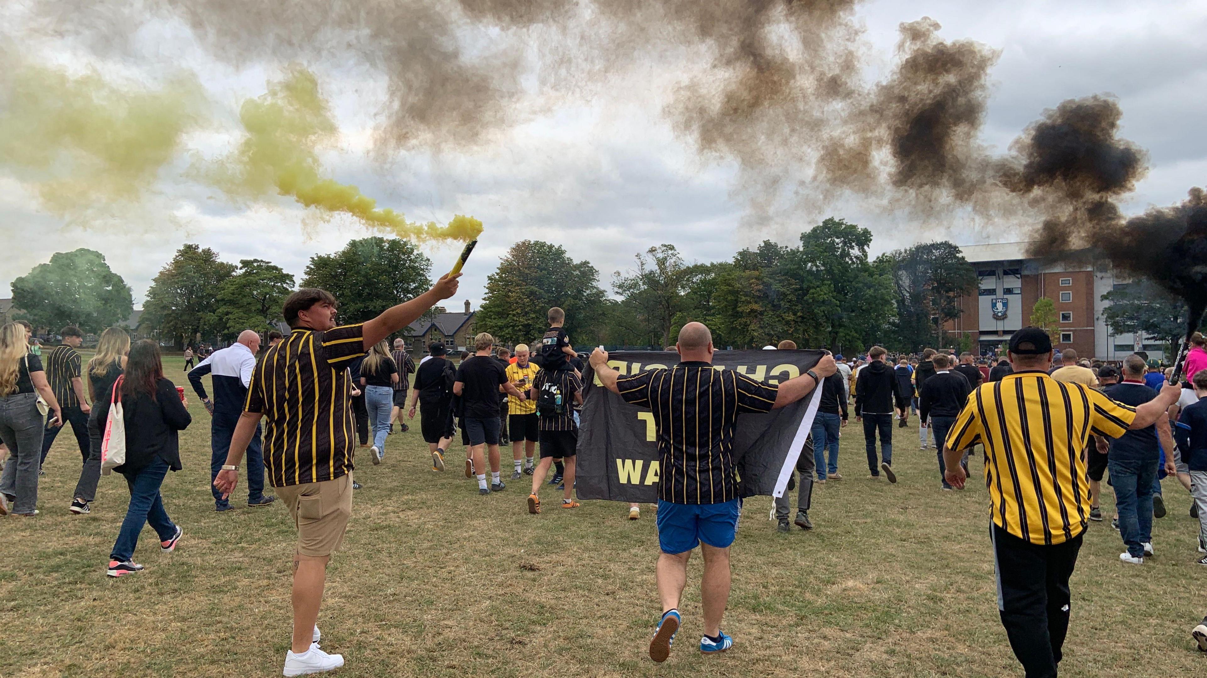 A group of people are walking through a park, some are wearing black and yellow striped Sheffield Wednesday shirts. One man has a yellow flare in the air and another man has a black flare