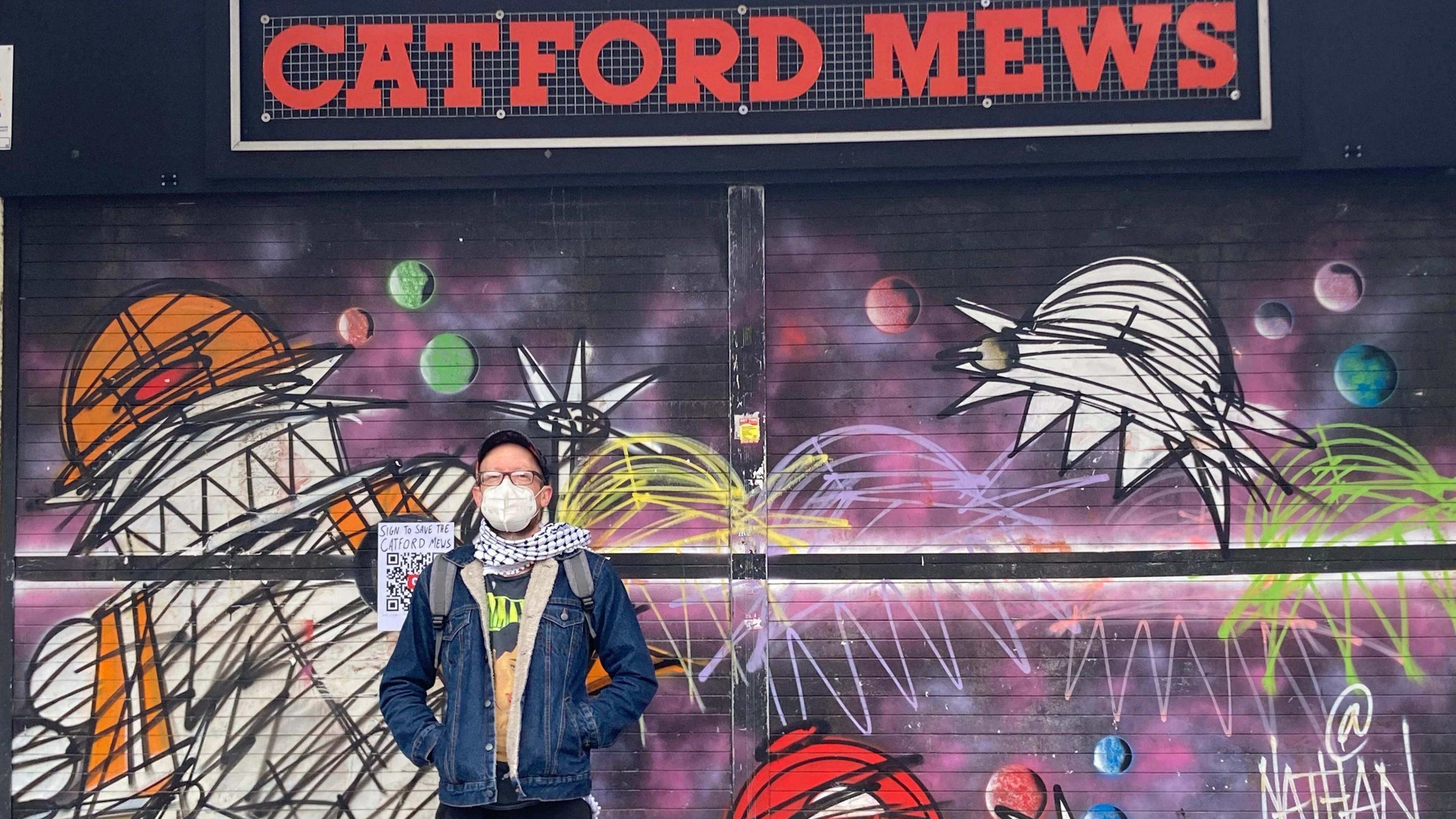 Zak Kilburn is wearing a mask, cap and denim jacket with a black and white scarf. He is stood facing the camera with the Catford Mews behind him. The shutters are covered in space-themed graffiti. 