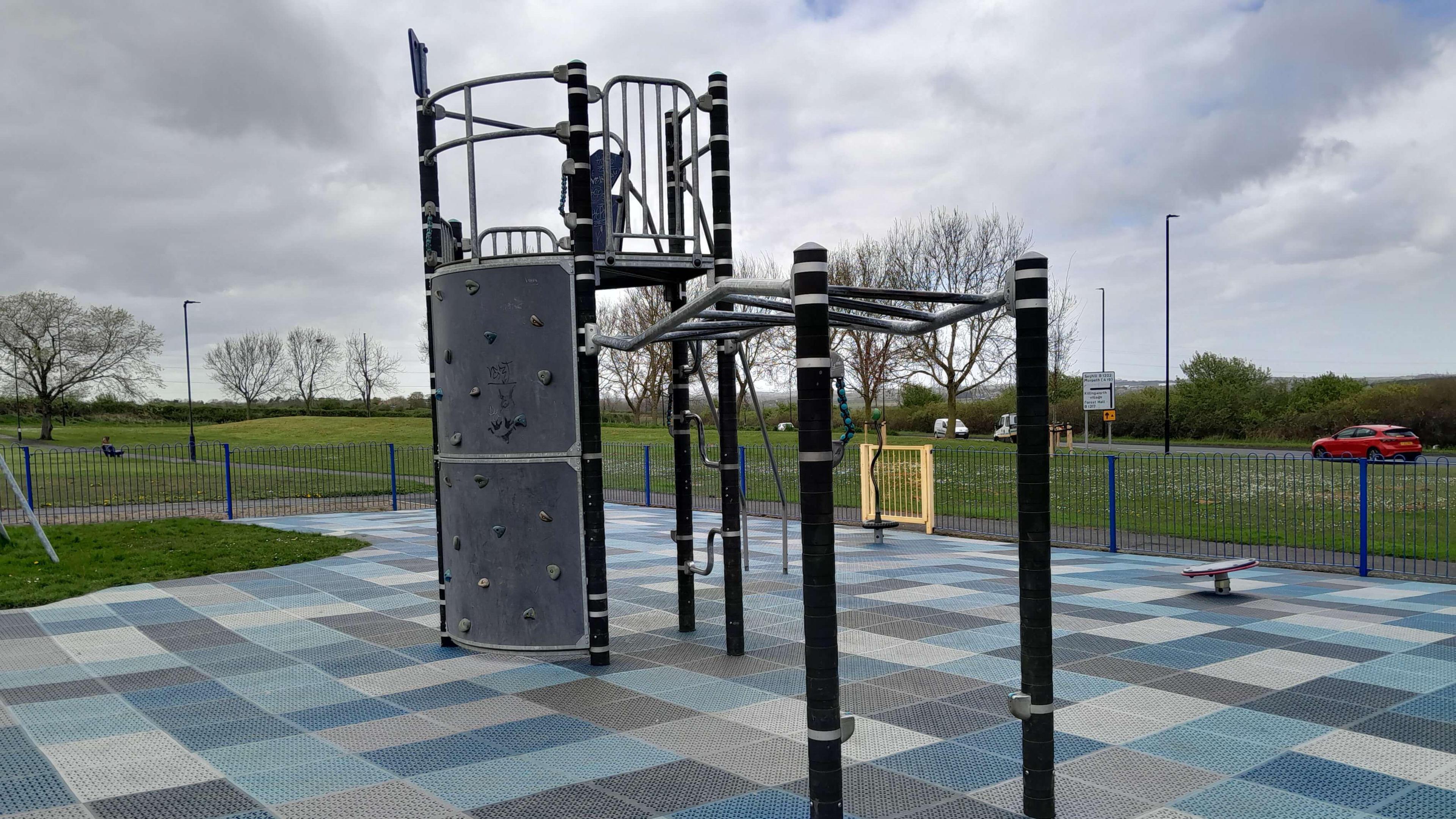 Blue and grey checkboard type rubber paving under a large climbing frame. There's blue fencing around it and grass in the background.
