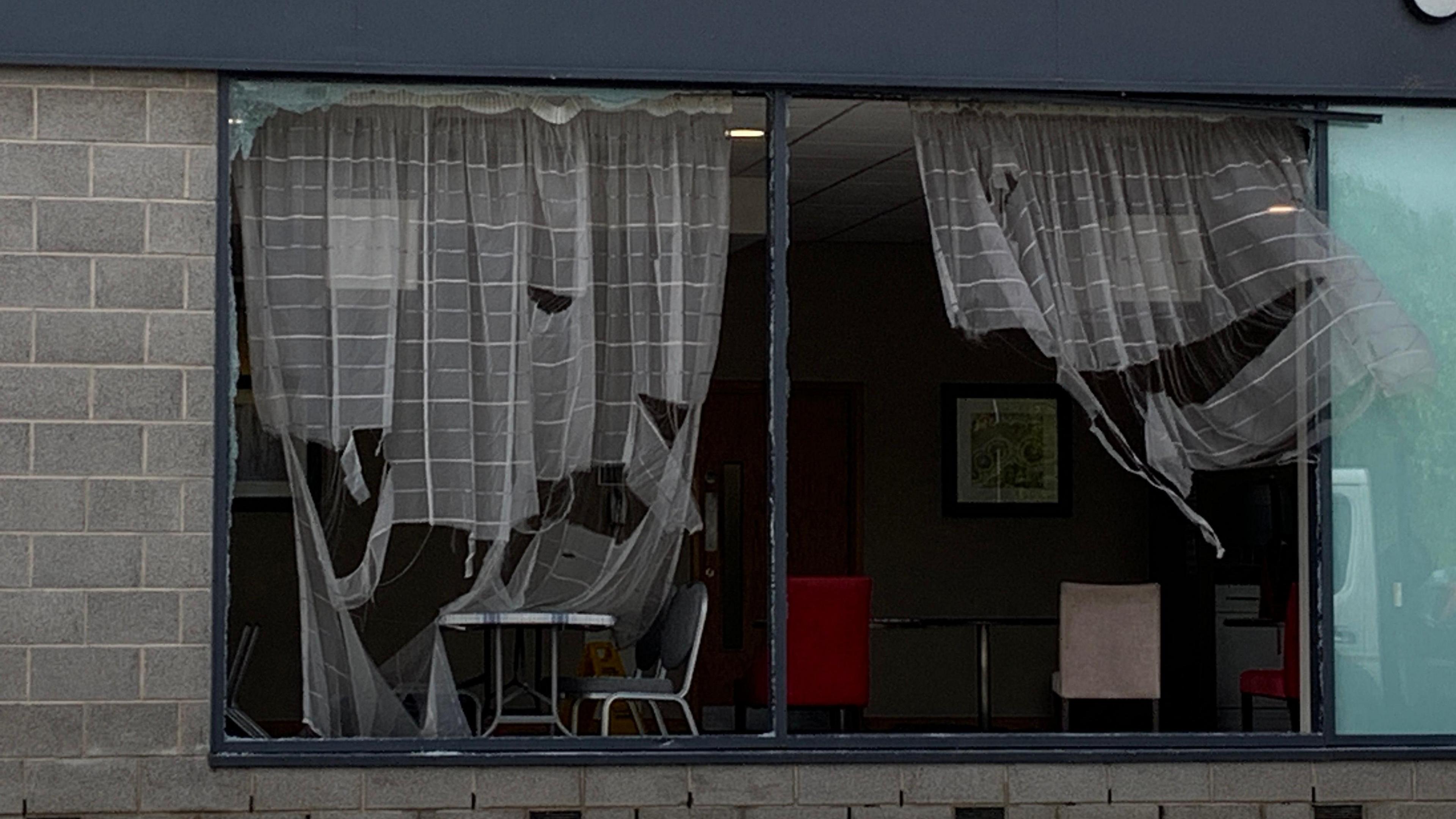 A picture of a window front. The glass is missing and ripped curtains can be seen hanging in the window. Several chairs and tables can be seen in front of the window.