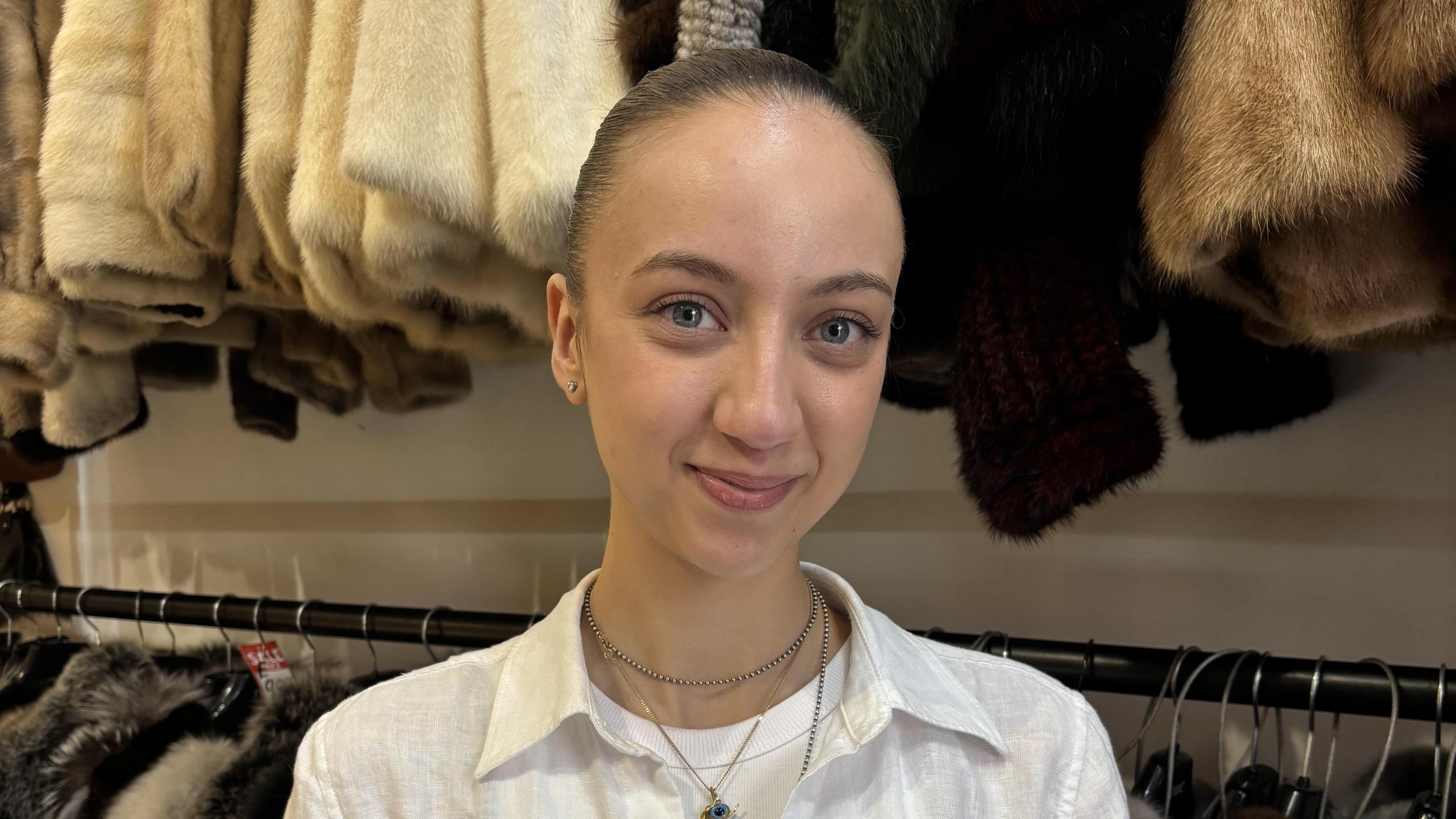 Mel Kaplan smiles at the camera. Visible behind her is a rack of fur coats in shades of white, cream, black and brown. Mel has her hair slicked-back into a ponytail and is wearing three layered necklaces and a satin shirt over a white T-shirt. It is a head and shoulders shot of her.