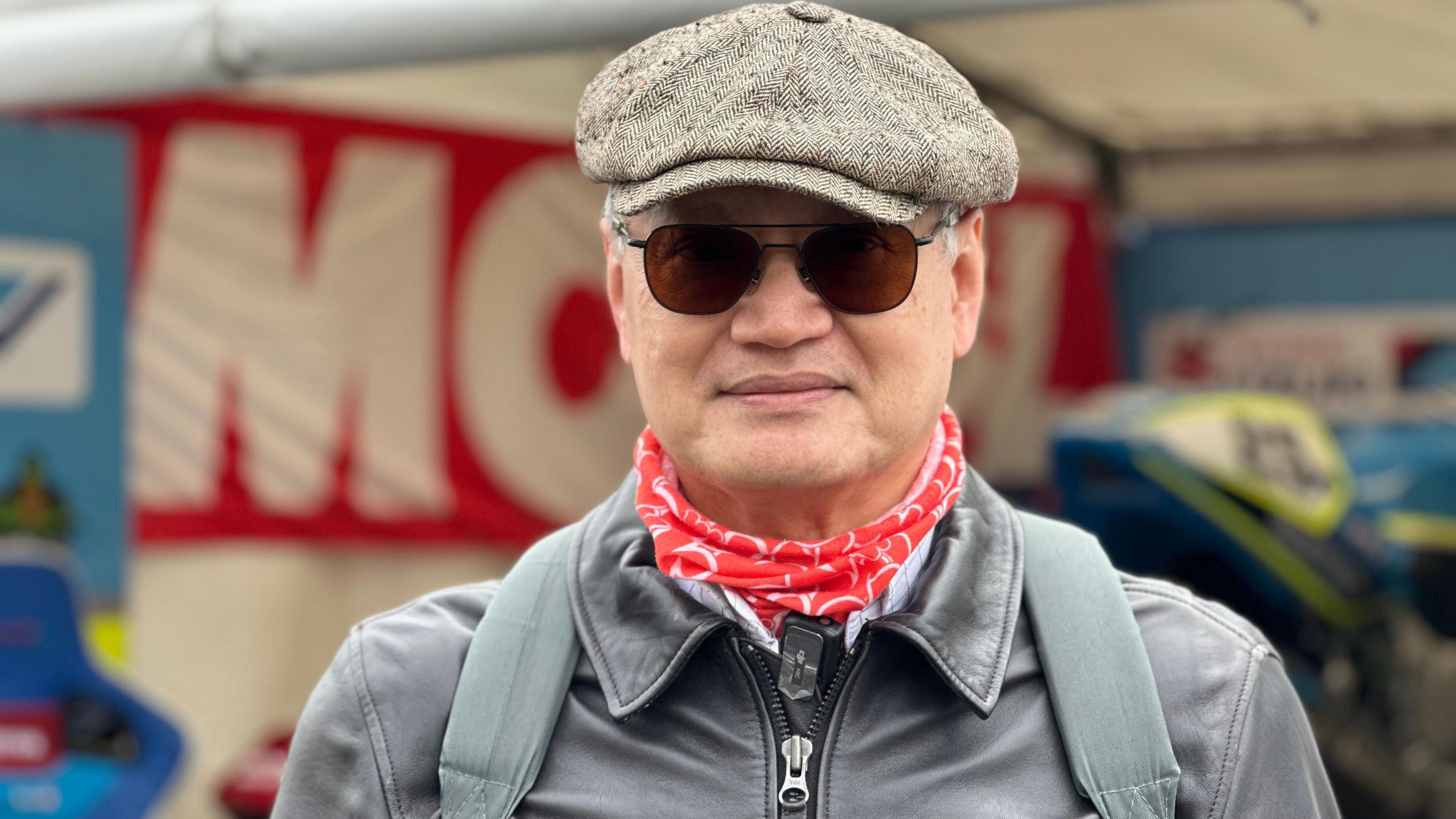 James Yeo stands in front of a tent that has a bike in it, which is out of focus in the background. He wears a tweed flat cap, sunglasses, a red scarf and black leather jacket.