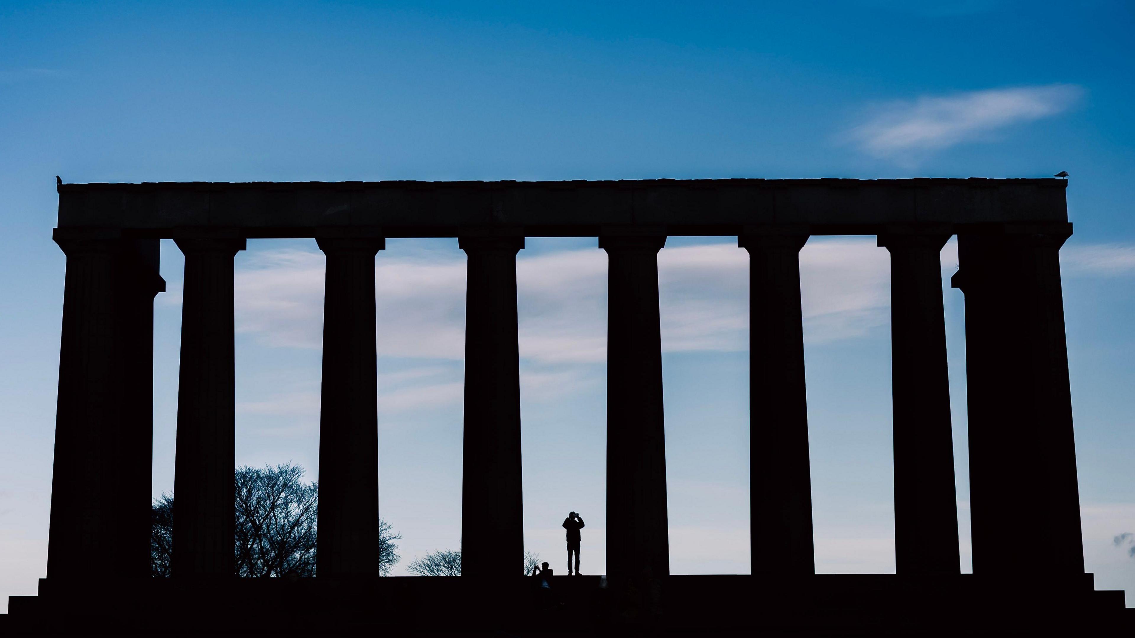 The silhouette of a person standing in between a column in front of a picturesque blue sky