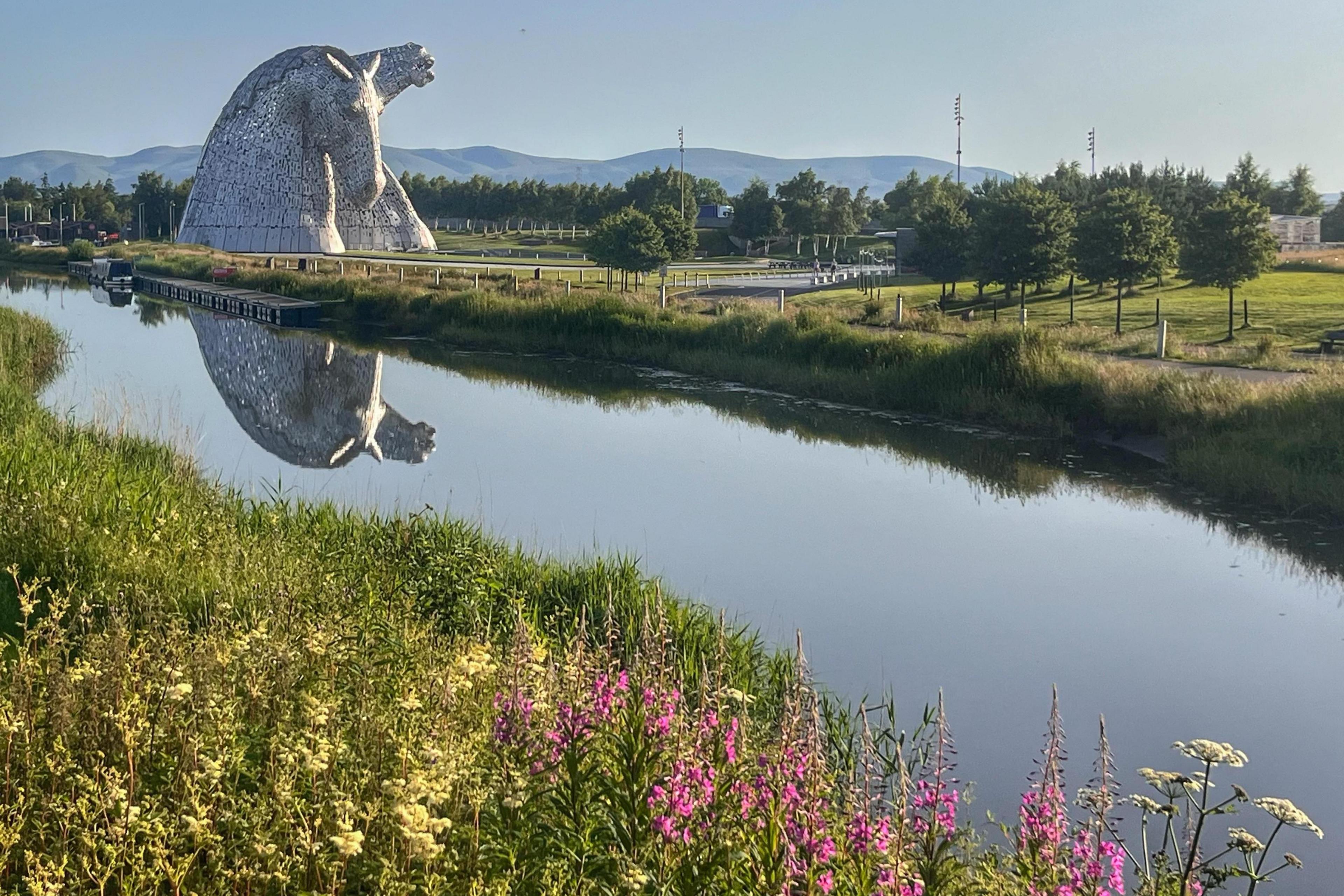 Large steel horse-head sculpture beside a calm canal, its reflection mirrored in the water, with green parkland, trees, and distant hills under a clear blue sky.