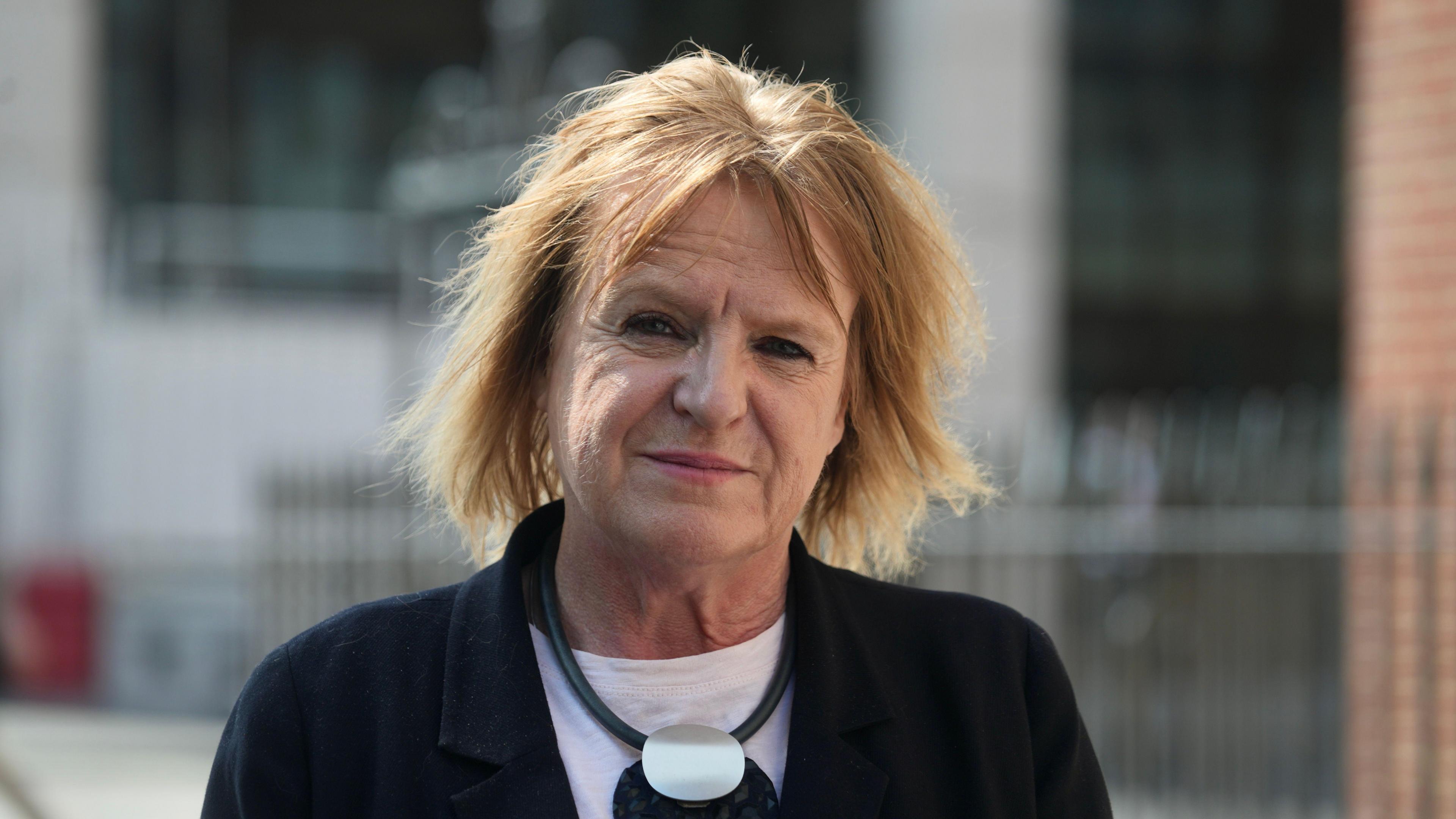 Head and shoulders photo of Deborah Coles. She has straight blonde shoulder-length hair with a centre parting. She is wearing a dark jacket with a white top underneath, and a large black and silver necklace. She is standing in front of a building with railings outside, and is looking directly into the camera