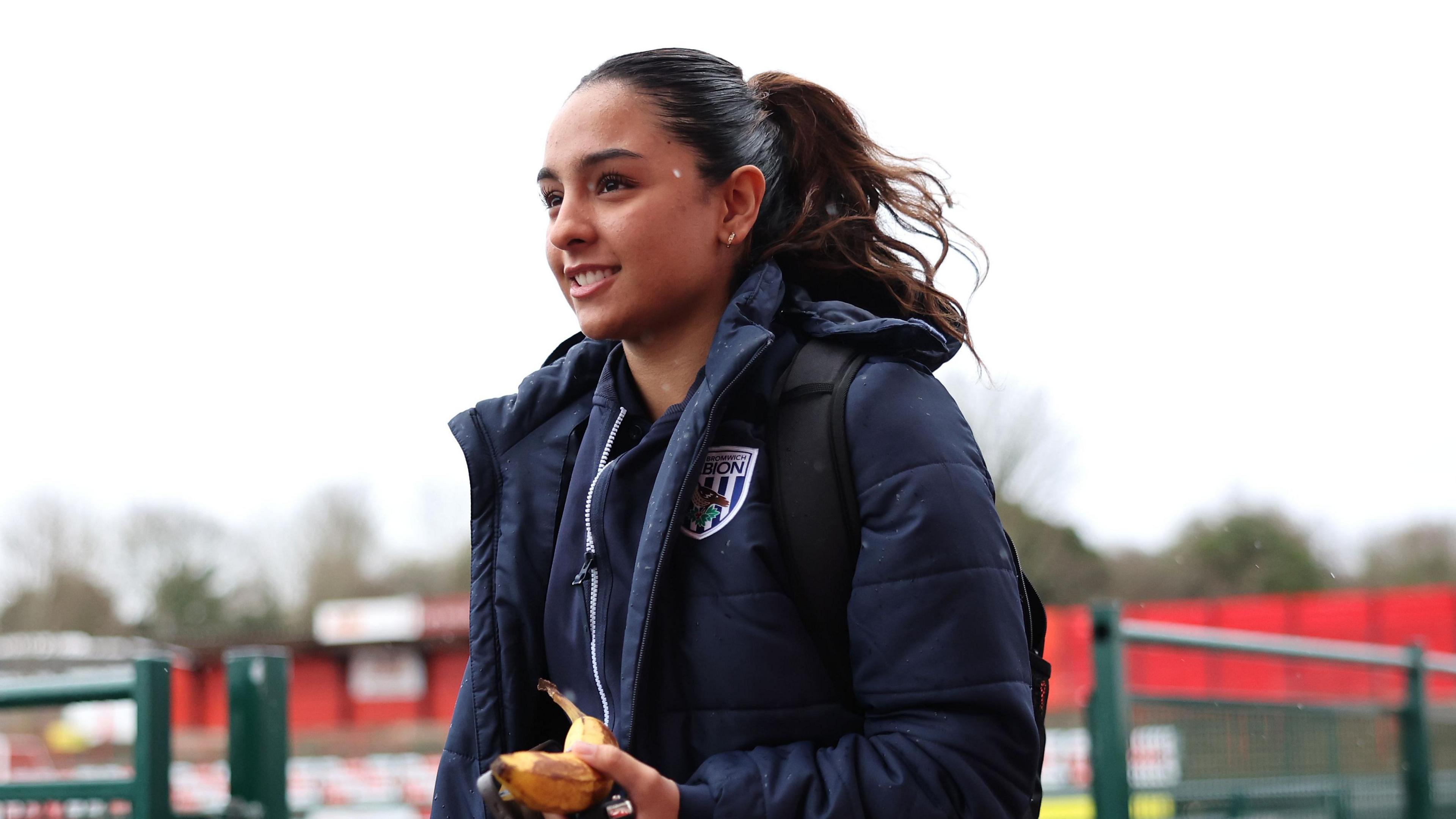 Mariam is smiling with a banana in her hand. She has a navy winter jacket on. In the background you can see a football pitch.