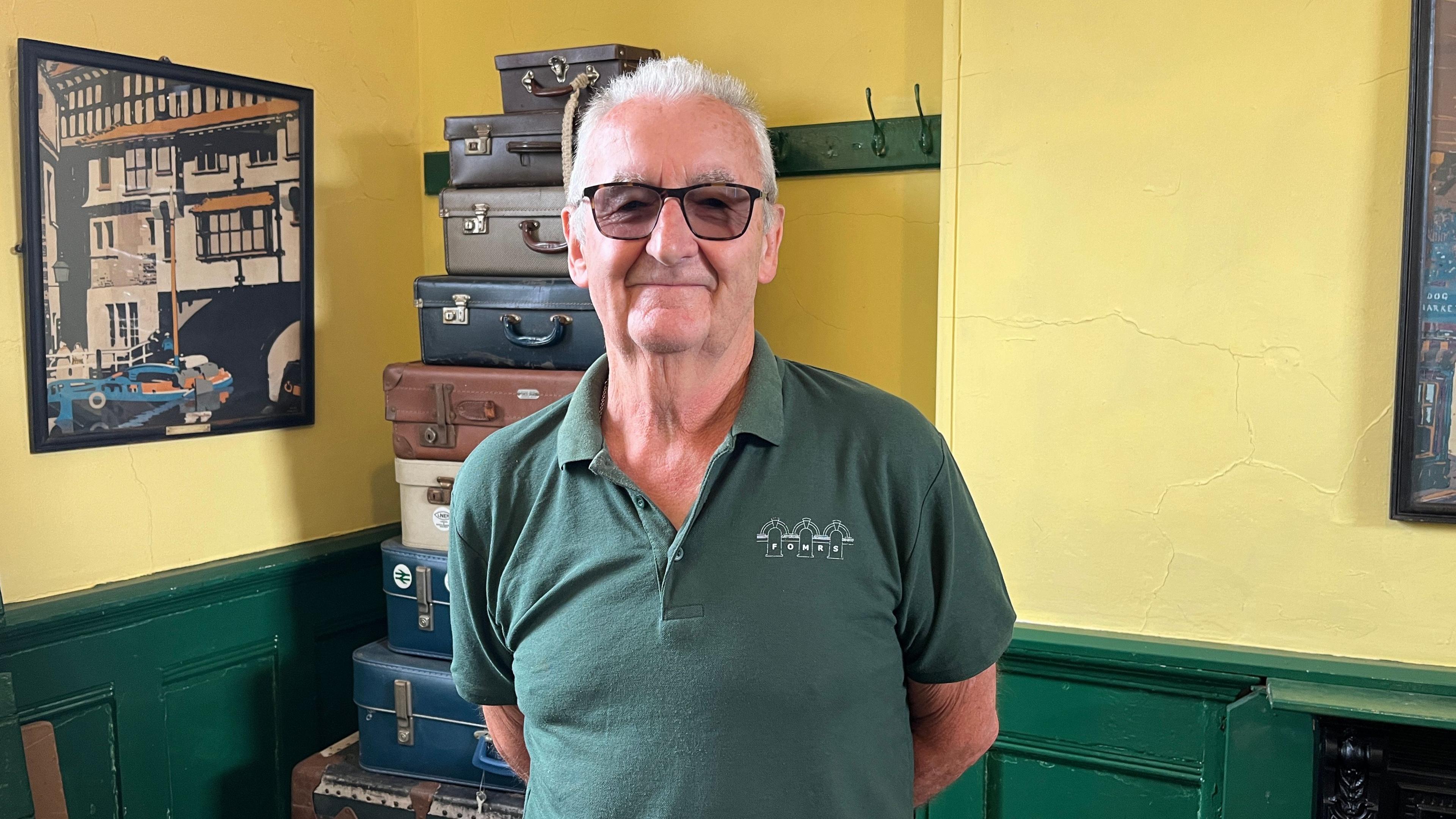 Volunteer and librarian Dave Story stands in the heritage room, a recreation of a Victorian waiting room. He has short grey hair and is wearing sunglasses and a green polo shirt. Behind him is a stack of vintage suitcases. There is an old poster on the wall showing a boat going under a bridge, and the corner of a cast iron fireplace can also be seen. The room is painted yellow and green.