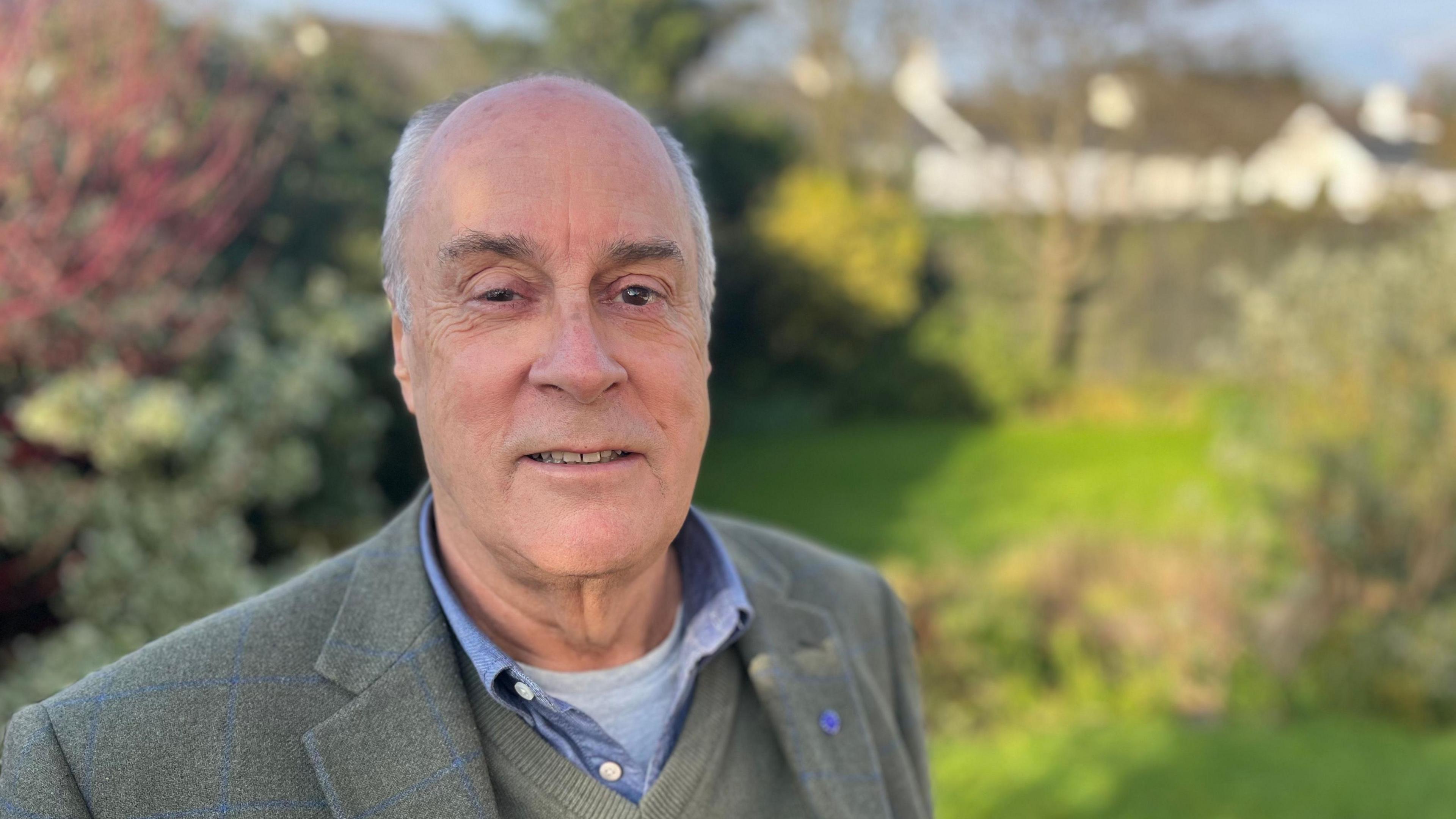 Liberal Democrat councillor Alistair Dewhirst wearing a blue shirt, olive sweater and an olive blazer against a blurred backdrop of garden