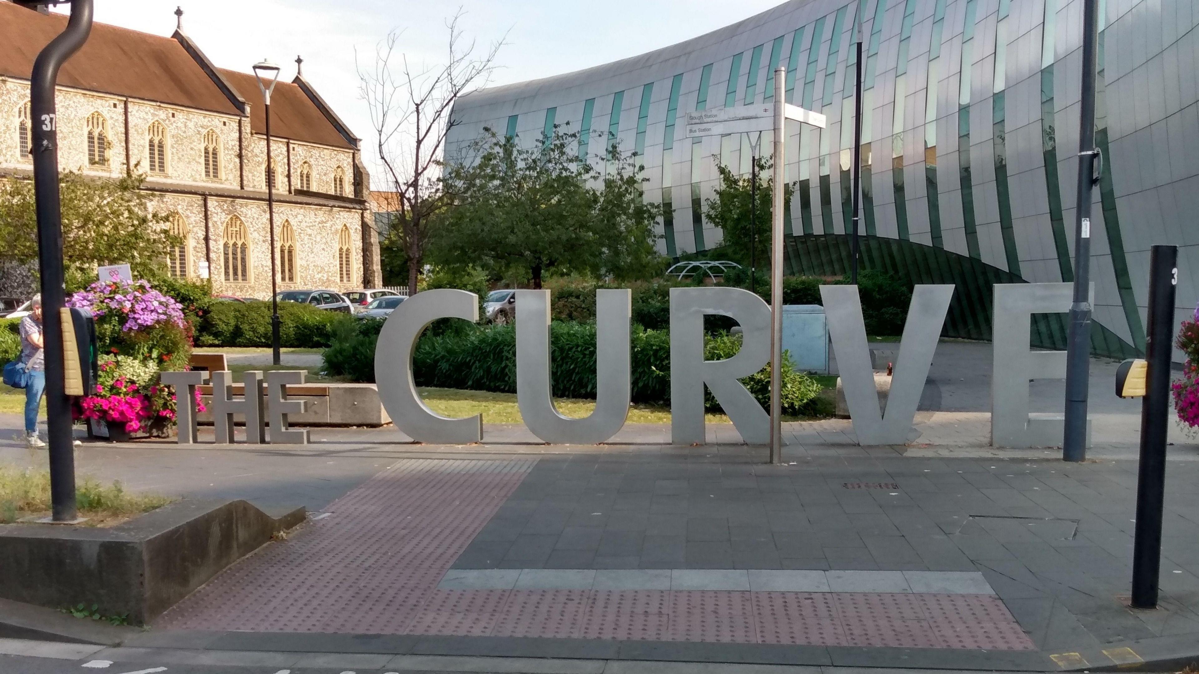 Big concrete letters, about the height of a person, saying 'The Curve' stand on a street in front of a pedestrian crossing. Behind the letters is a large silver building with a curved wall.