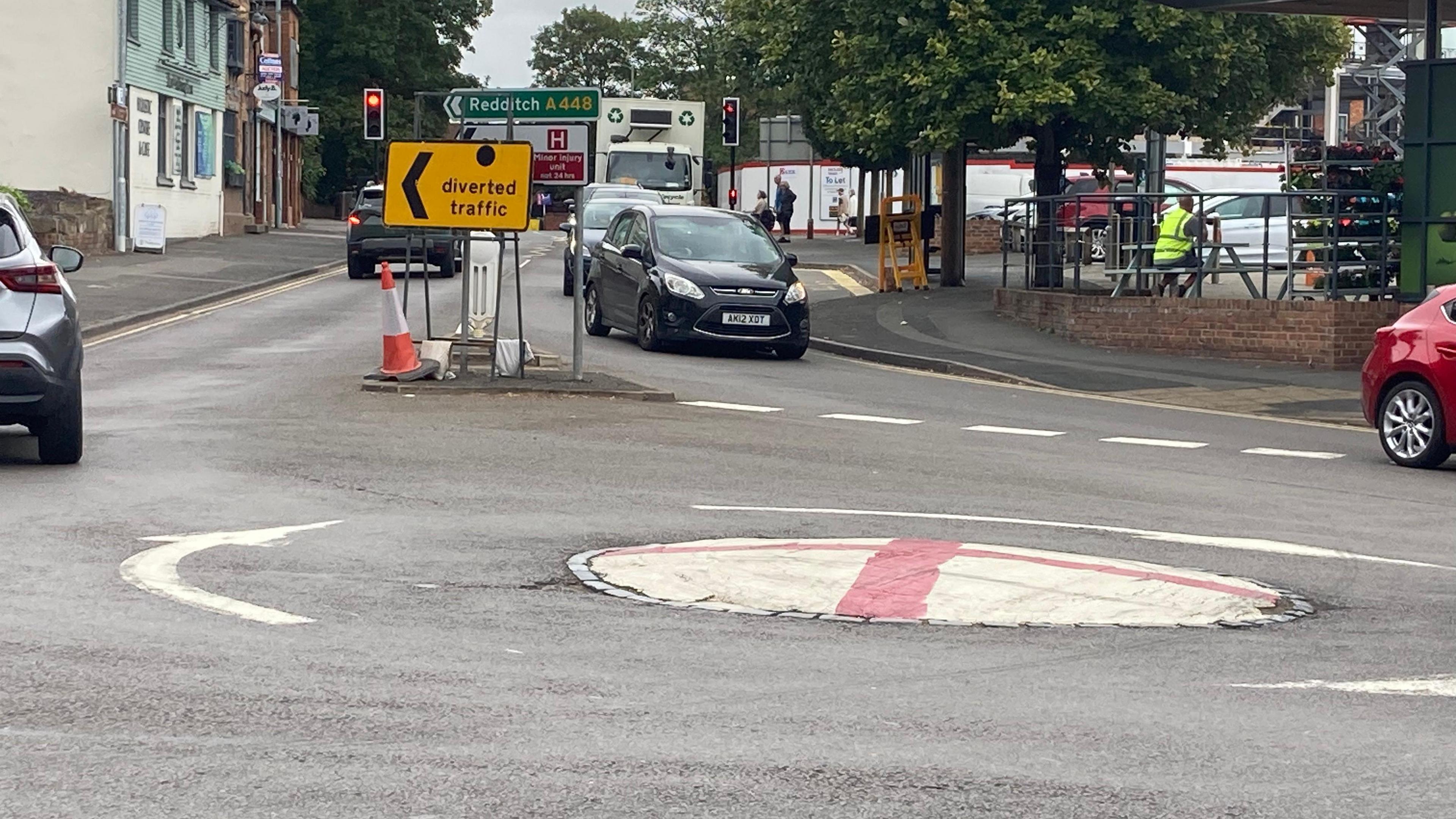 A mini-roundabout painted with the flag of St George - with cars going in two directions and a road sign showing Redditch. 
