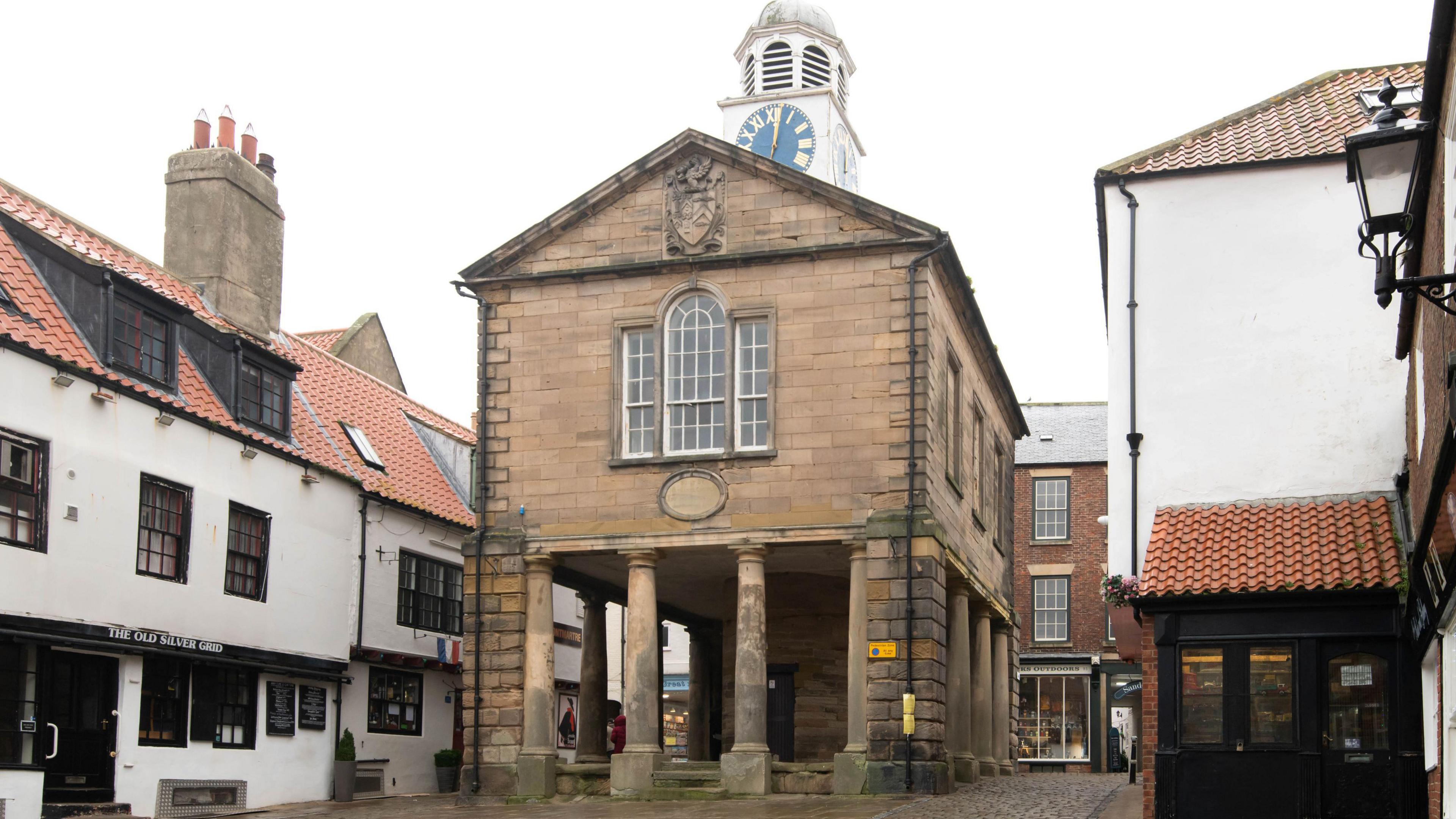 A frontal view of Whitby Old Town Hall, which features pillars, an under croft, yellow bricks, and a black clock face on a white tower above its roof.