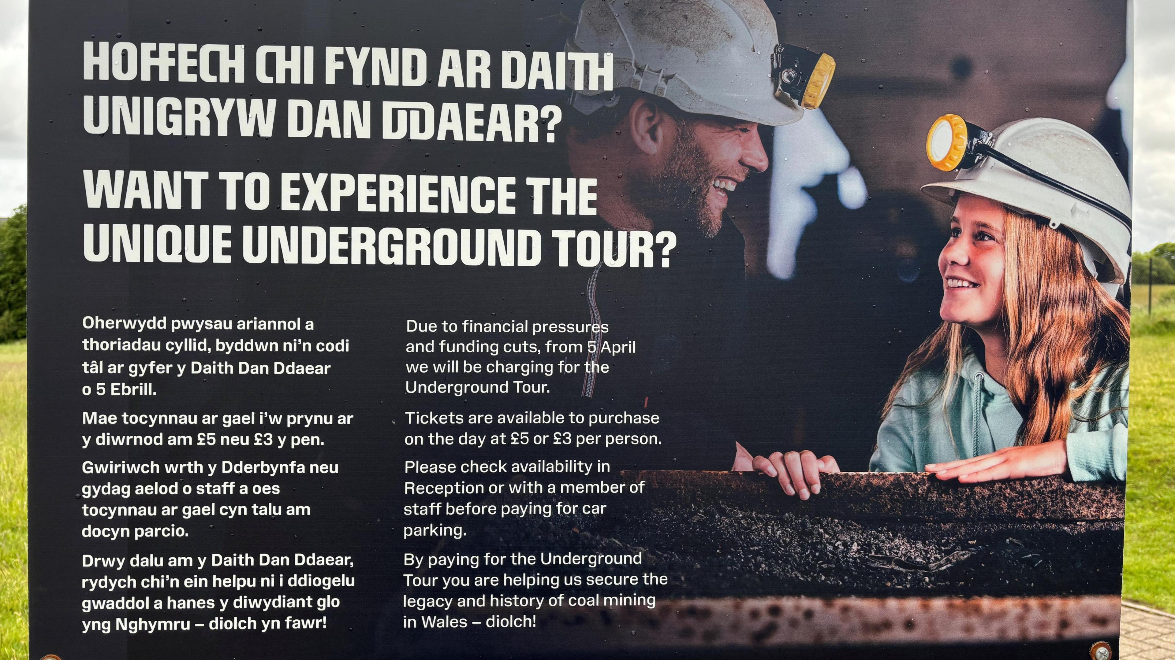 A sign telling visitors that Amgueddfa Cymru will be charging for underground tours at the Big Pit Musuem due to financial pressures. The sign has a background picture of a man and a young girl wearing white hard hats in an underground tour
