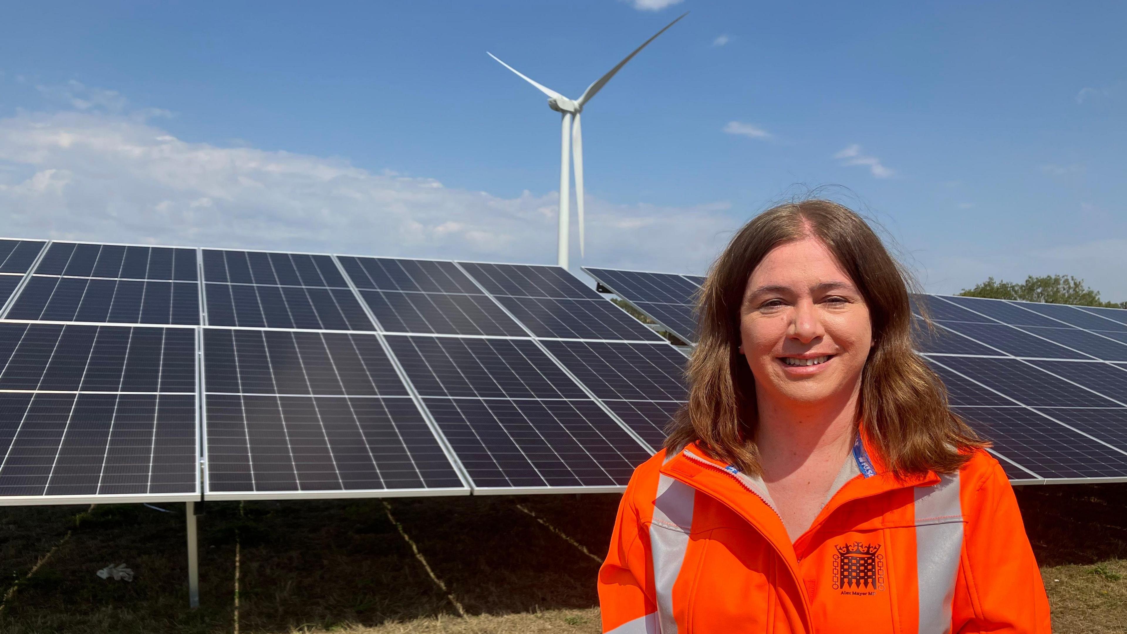 A picture of Labour MP Alex Mayer, a woman with shoulder length dark brown hair in a centre parting. She is wearing a high visibility jacket and is stood in front of some solar panels.