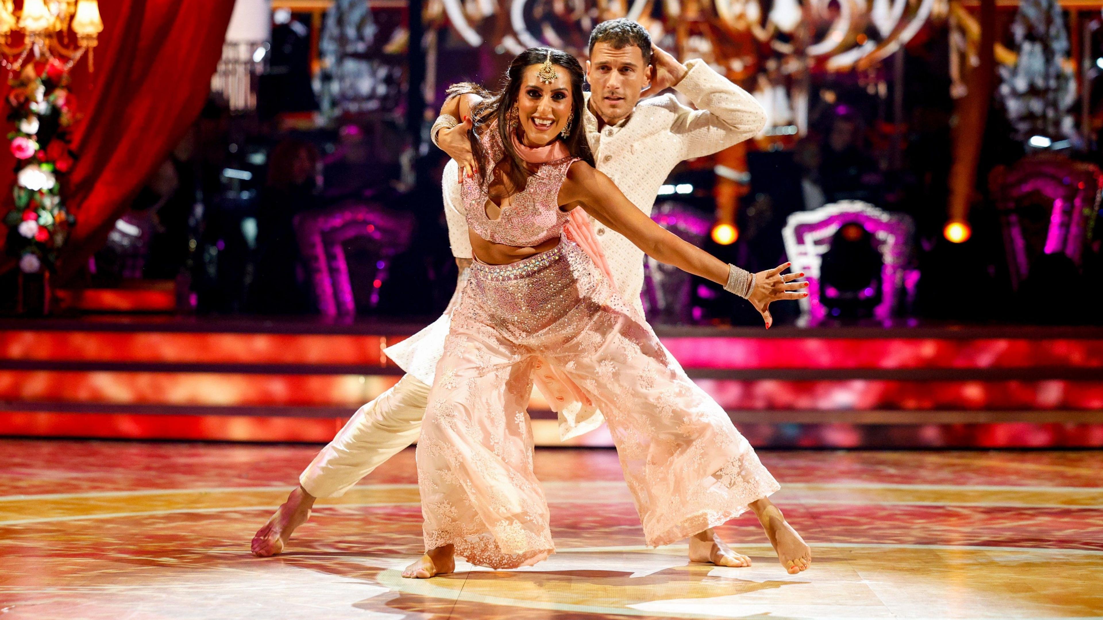 Dr Punam Krishan mid dance on the Strictly dancefloor, she is wearing a pink jewelled traditional Bollywood costume. She has her left arm and right leg extended. Her dancing partner Gorka Marquez stands behind her wearing a white Bollywood outfit.