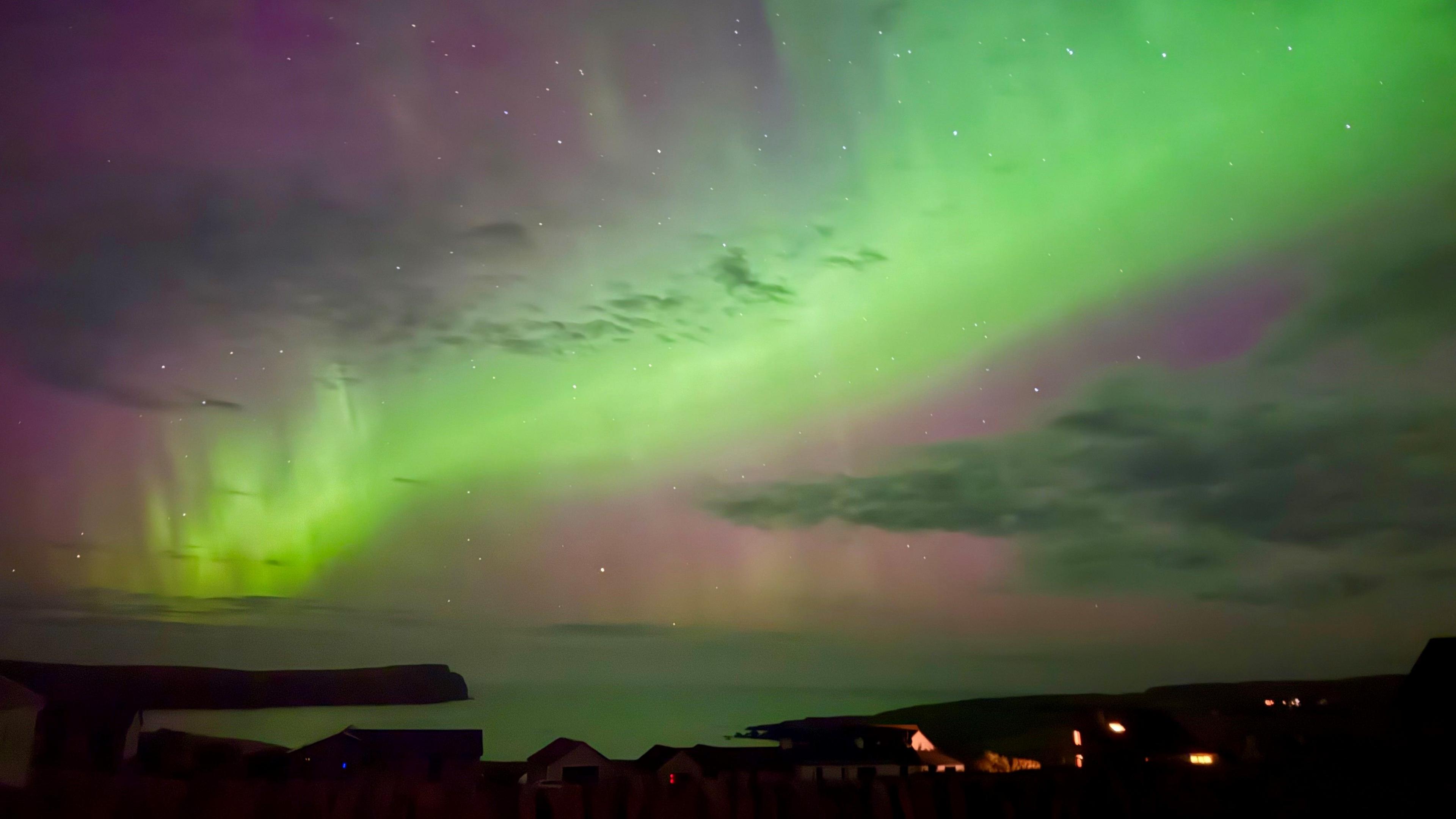 A coastline dotted with lights from houses are in the foreground with the sea stretching out into the distance with the Northern Lights above.