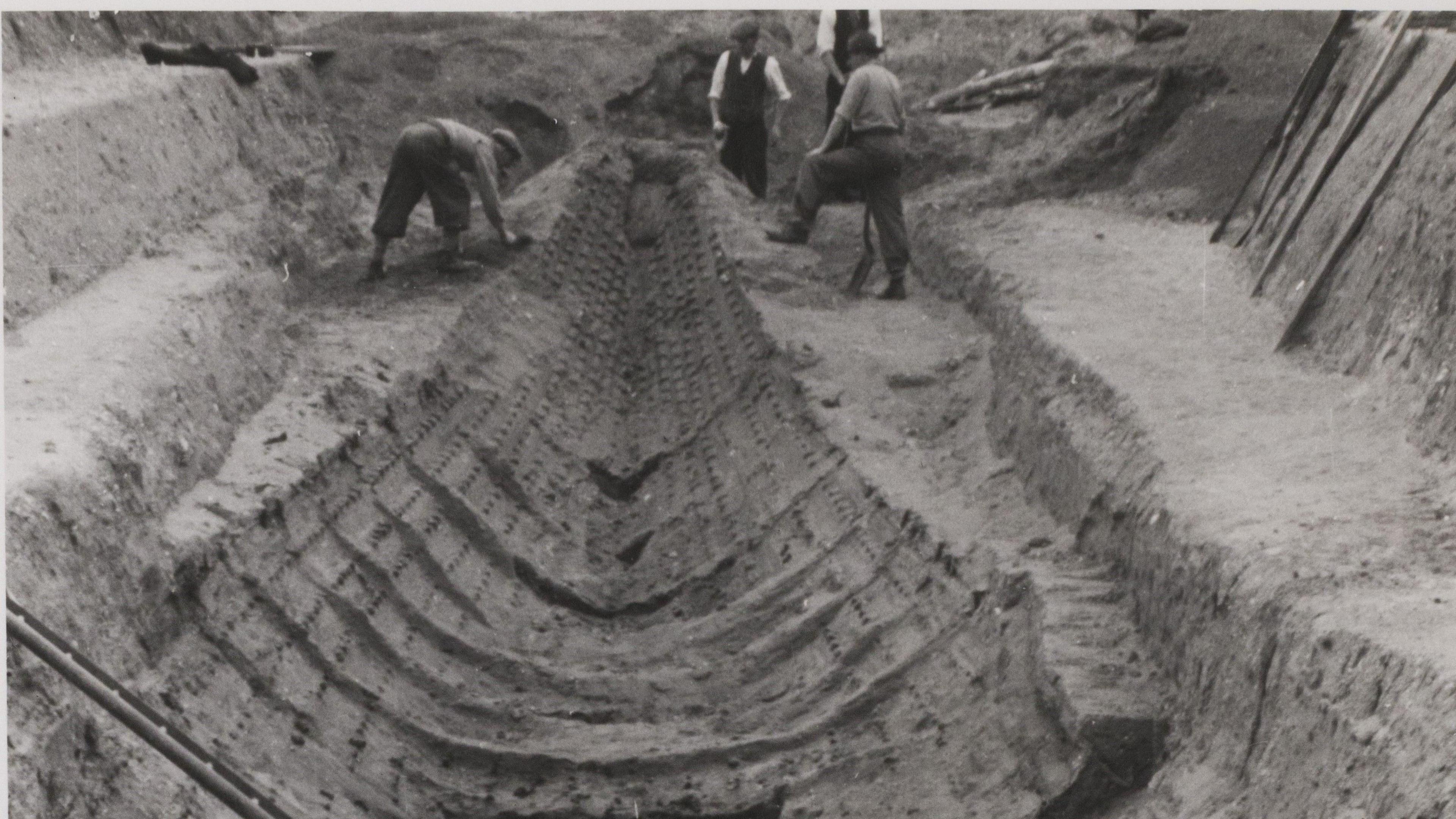 The original burial sheep unearthed at Sutton Hoo. It is a black and white picture and shows the hull of the ship being excavated.