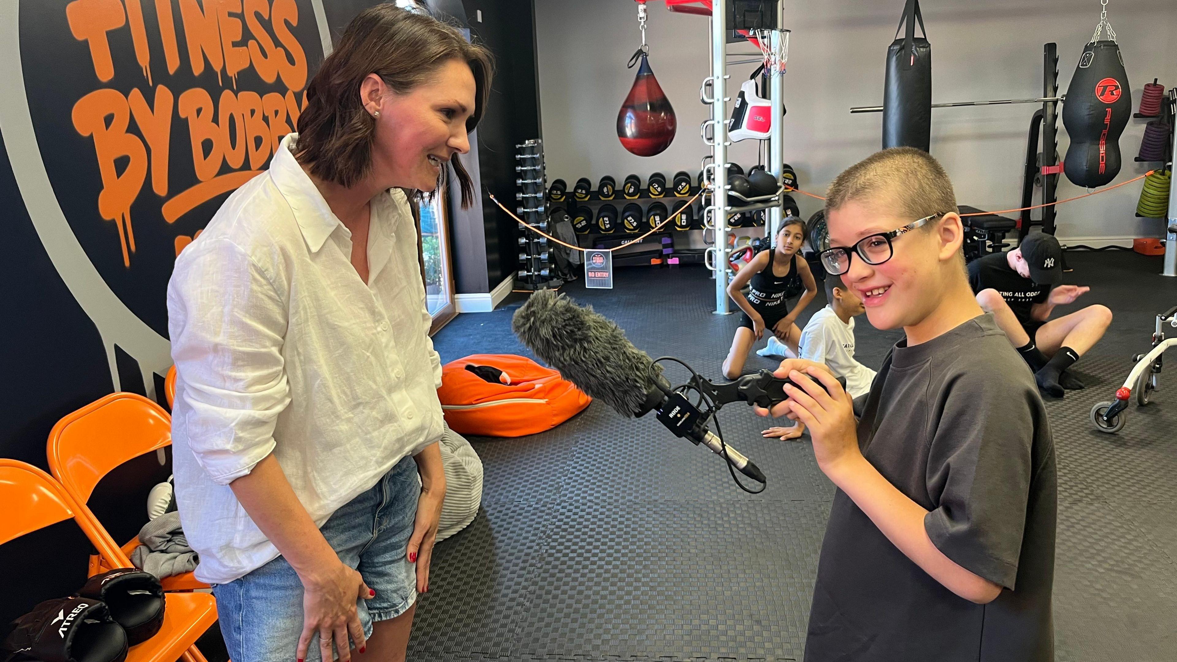 Mum Hayley is wearing a white short and demin shorts and is with her son Rex who is wearing a grey t-shirt. He has glasses and is holding a microphone.