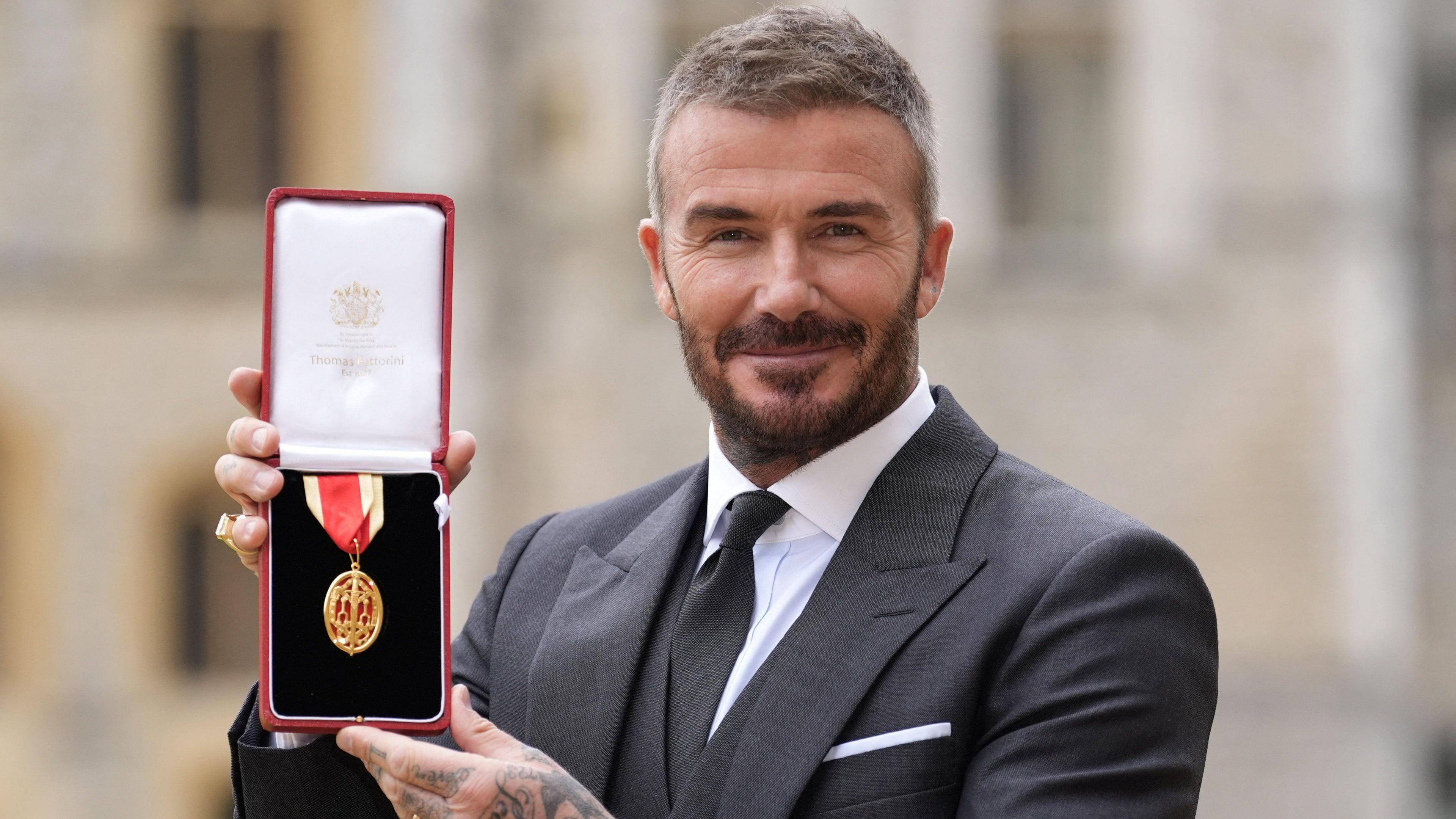 David Beckham holds his medal and smiles at the camera. The medal is red and gold and is in a box. Beckham wears a black suit and white shirt with a black tie.