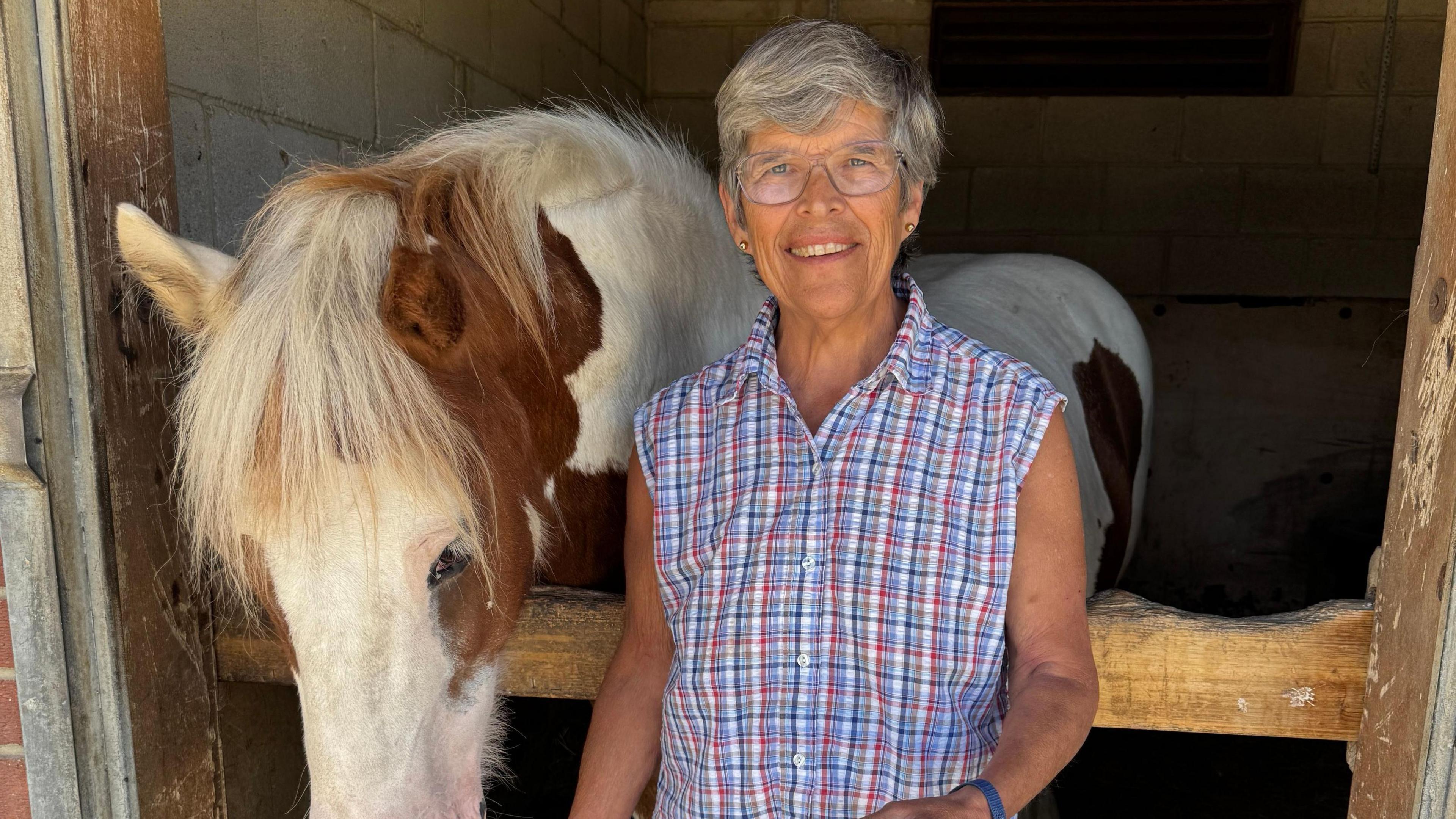 Deborah Hall is wearing a checkered, sleeveless shirt. She is standing next to a horse in stables.
