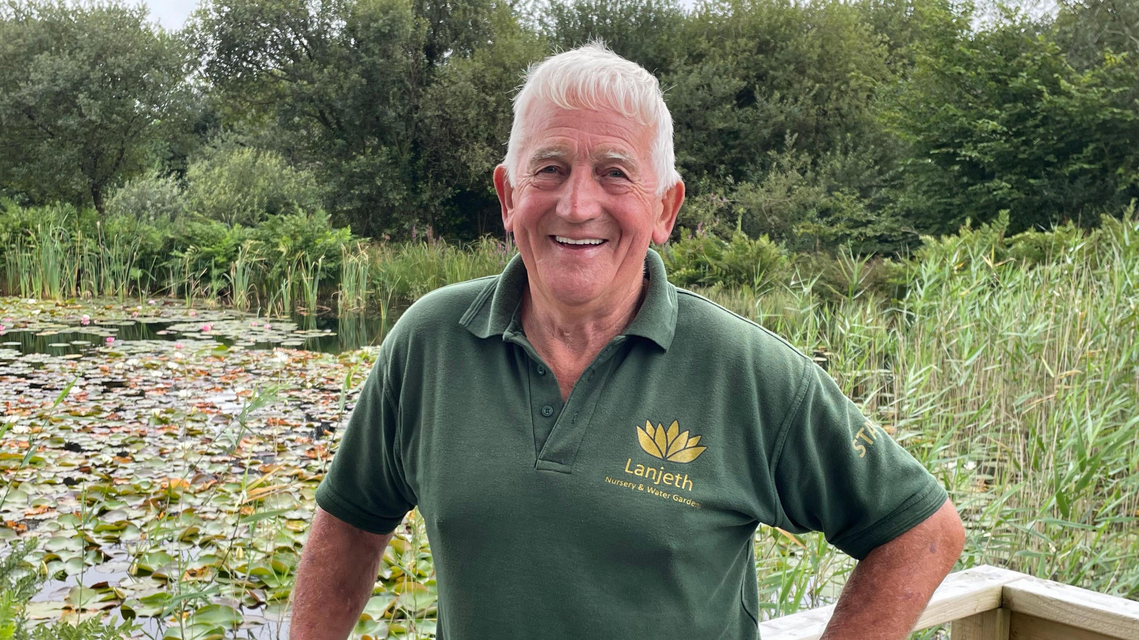 Norman Trebilcock is wearing a green polo shirt and is stood in front of a pond with water lilies and reeds