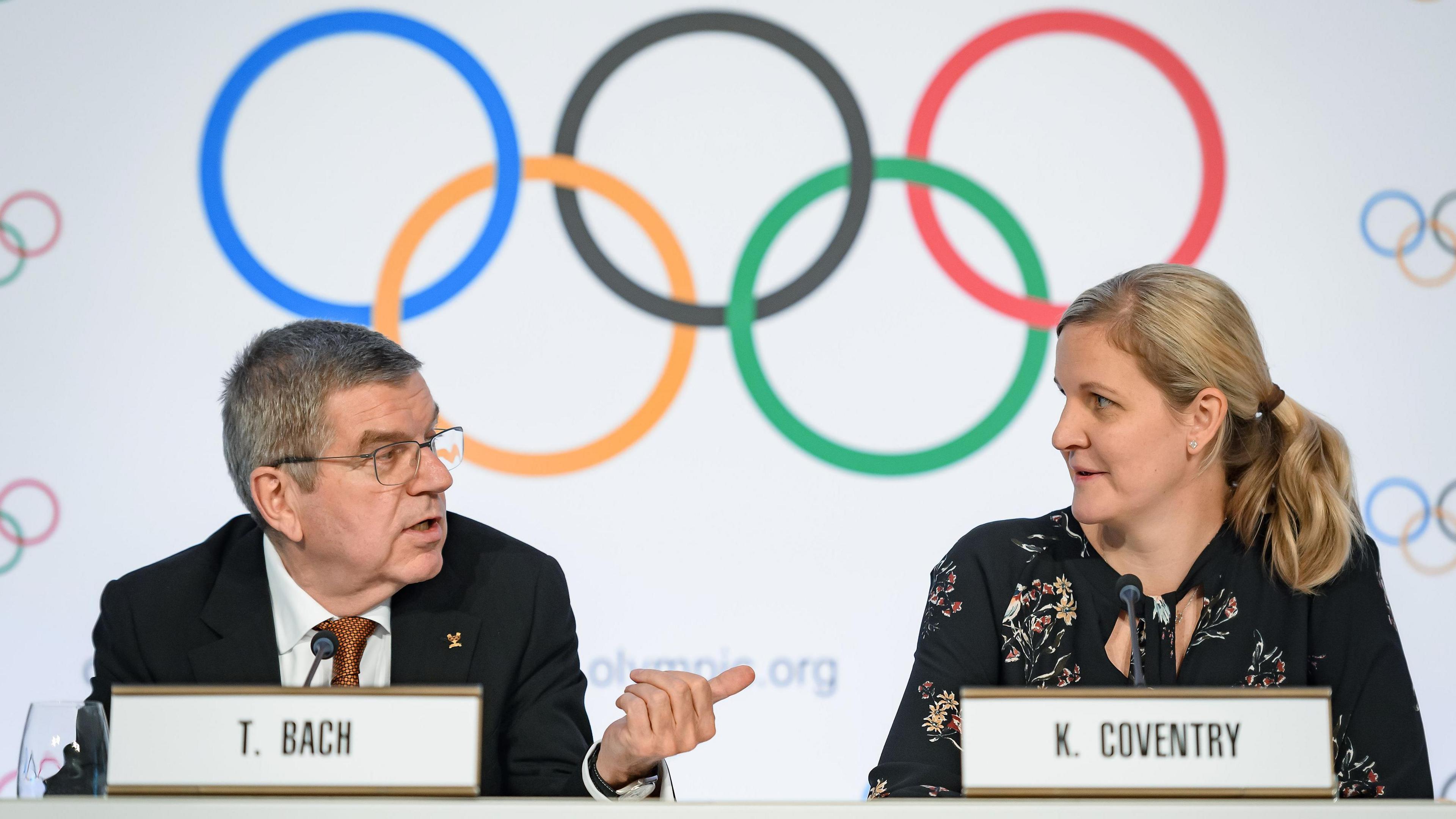 Seated at a table bearing nameplates with a background of the Olympic rings behind them, Tomas Bach looks to his left and points with his left thumb towards Kirsty Coventry